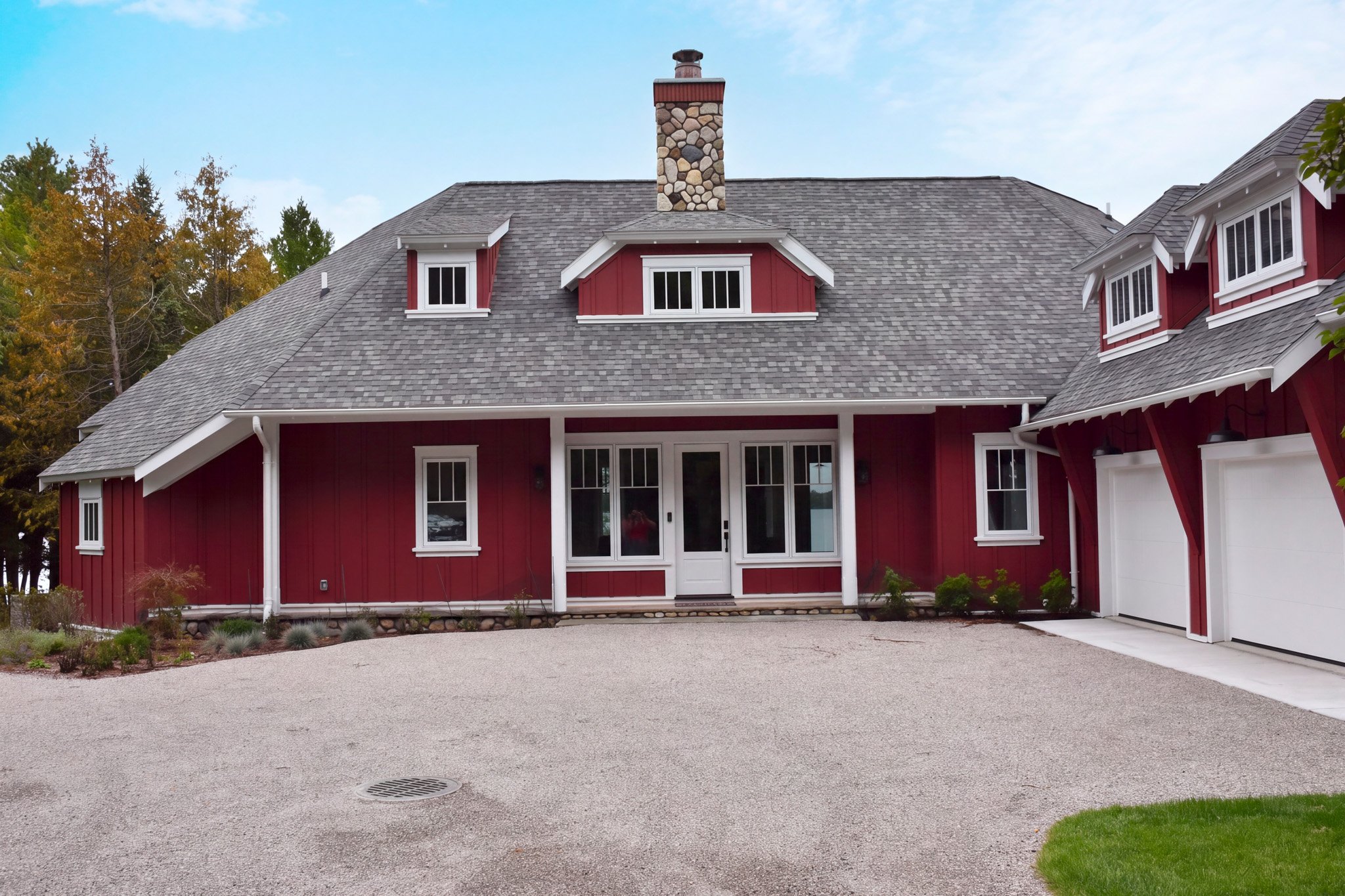 A red house with gray shingle roof, white trim, a stone chimney, several windows, and a white sliding door, surrounded by a gravel driveway and landscaped garden with trees and shrubs.