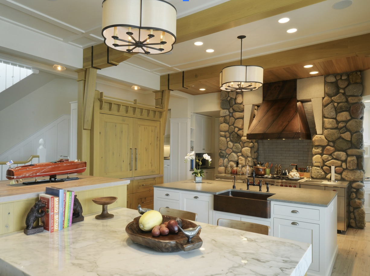 A spacious kitchen with a large island, white cabinets, a stone and wood fireplace, and modern lighting fixtures.