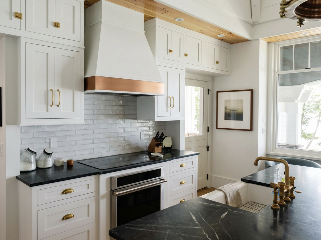 A modern kitchen with white cabinets, black countertops, a stainless steel oven, a white brick backsplash, and a copper range hood, with natural light coming through large windows.
