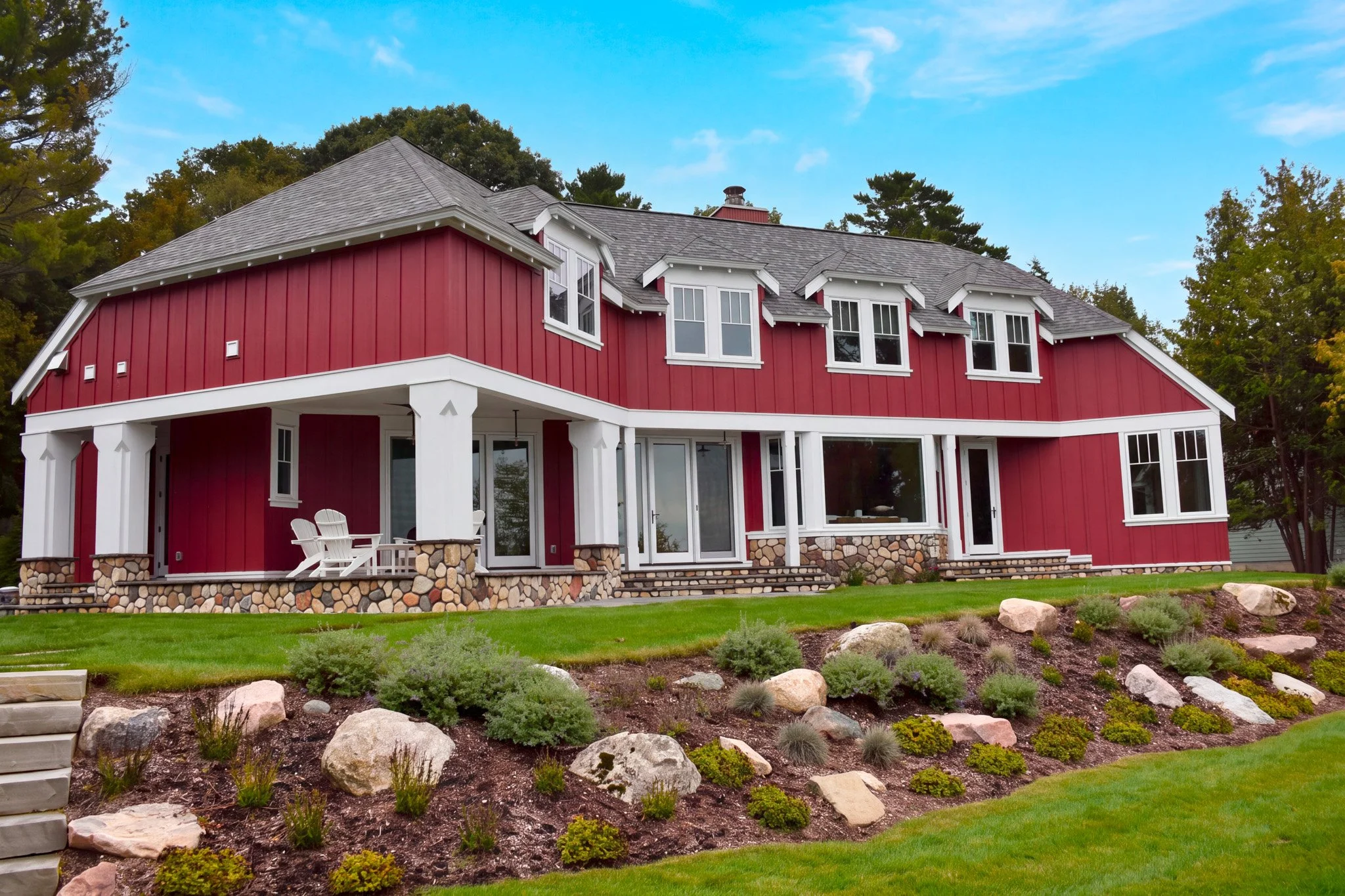 Red house with white trim and multiple windows, stone foundation, surrounded by landscaped yard with rocks and plants.