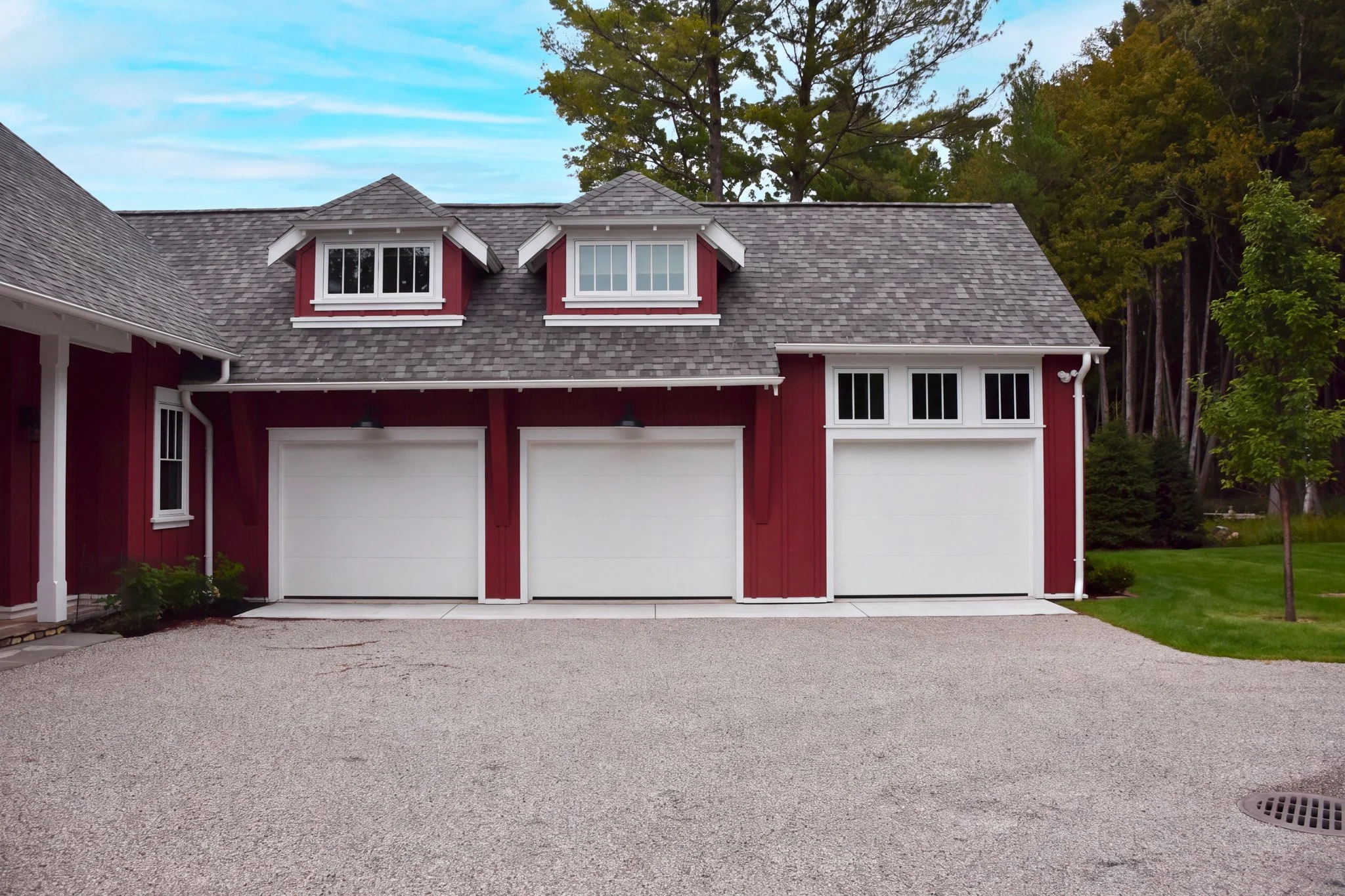 A modern house with a gray shingle roof, red exterior walls, and white trim, featuring two dormer windows and a garage. The driveway is made of gravel, and there are trees and greenery in the background.