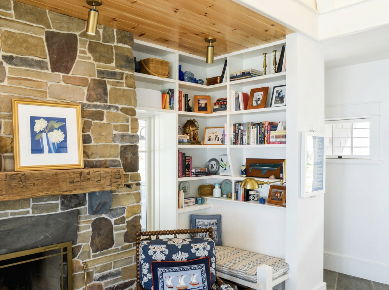 A cozy living room corner featuring a stone fireplace with a wooden mantel, a white bookshelf filled with books and decorative items, and a window allowing natural light.