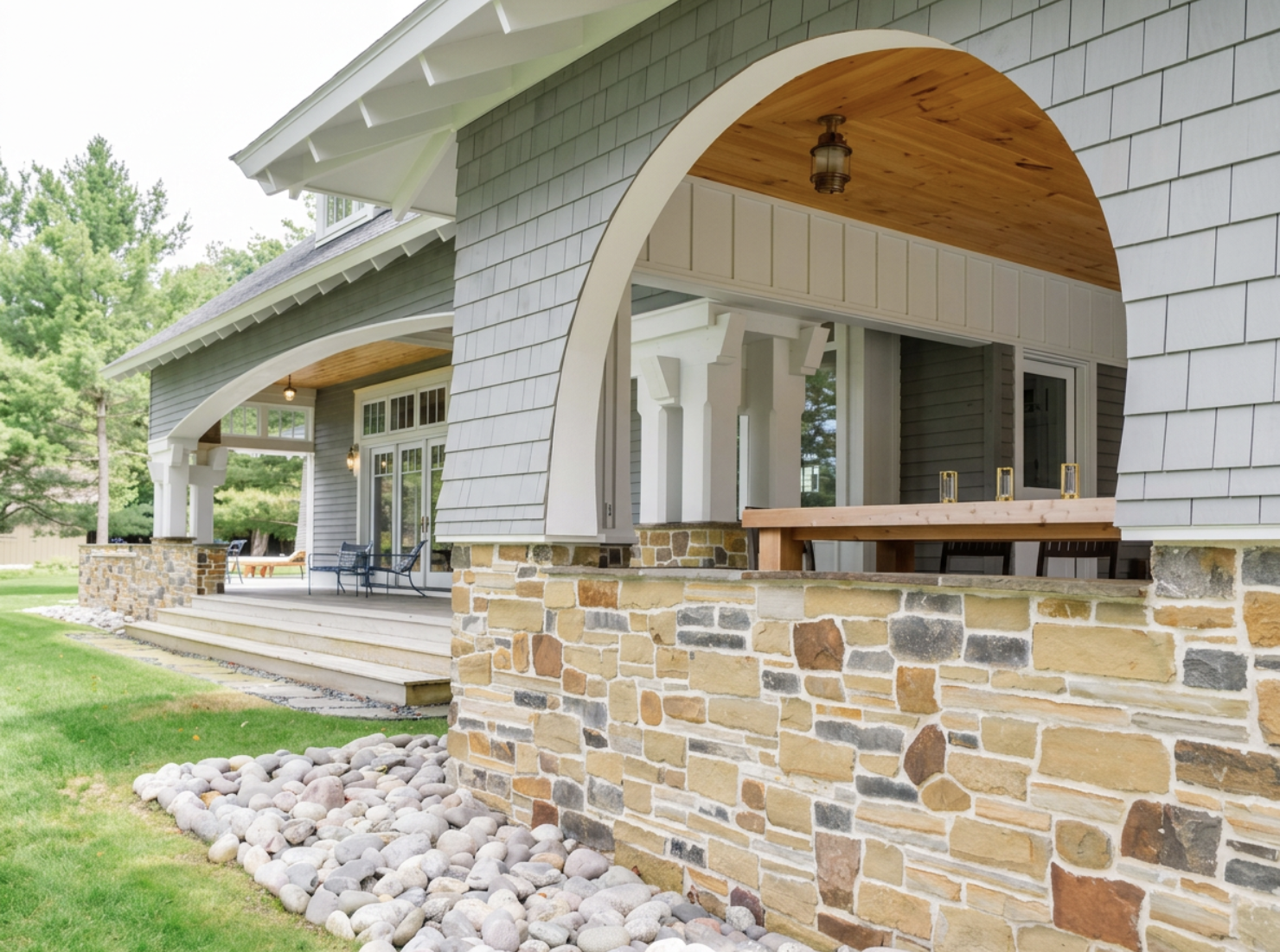 Porch with stone base, outdoor seating, arches, and a wooden ceiling, part of a house with gray siding, surrounded by green lawn and trees.