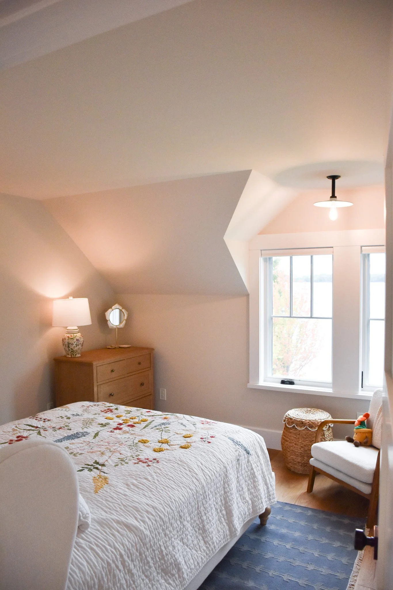 A cozy bedroom with a white quilted bedspread featuring floral embroidery, a wooden dresser with a floral lamp, a window with white blinds, a white armchair with a wicker side table, and a ceiling light fixture.