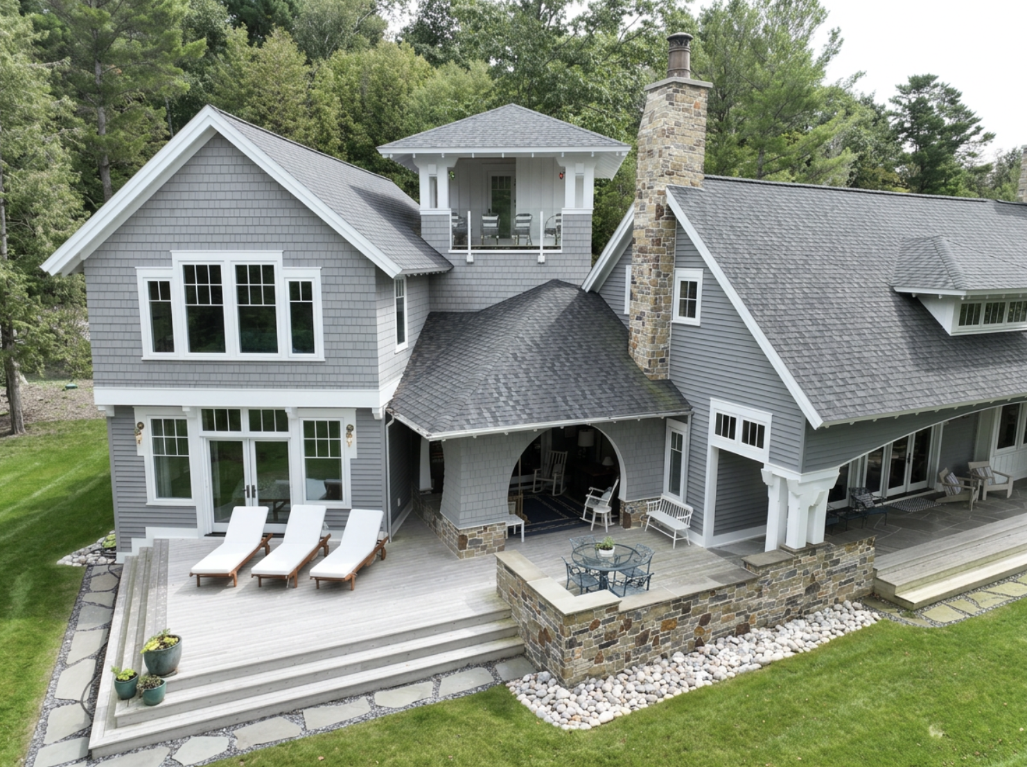 A large gray house with multiple sections, a stone chimney, and a spacious backyard deck with lounge chairs, outdoor dining table, and seating areas, surrounded by green grass and trees.