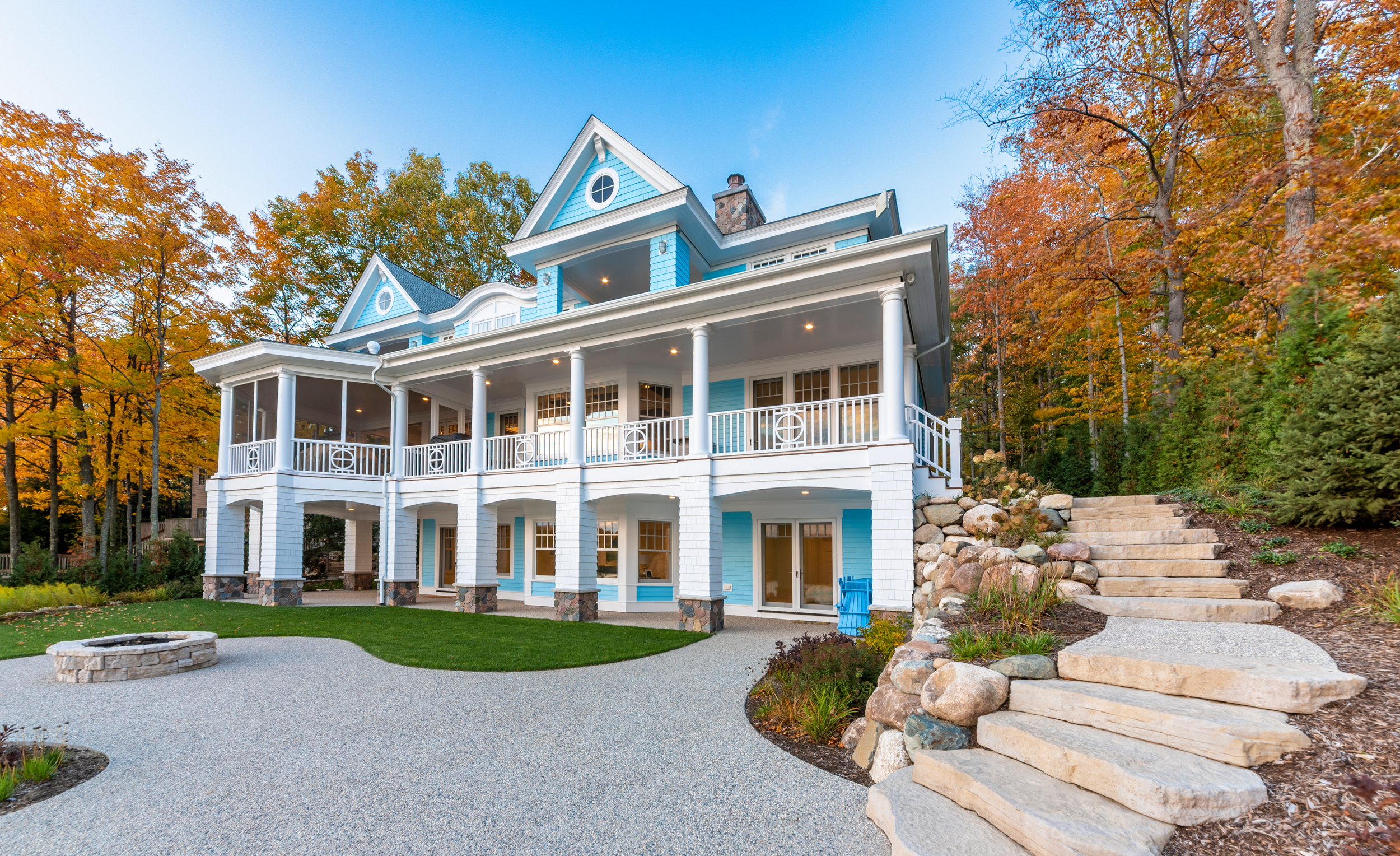 A large, multi-story house painted in light blue with white trim and a wrap-around porch, surrounded by fall foliage with orange and green leaves, a stone staircase on the right side, and a gravel path in the yard.