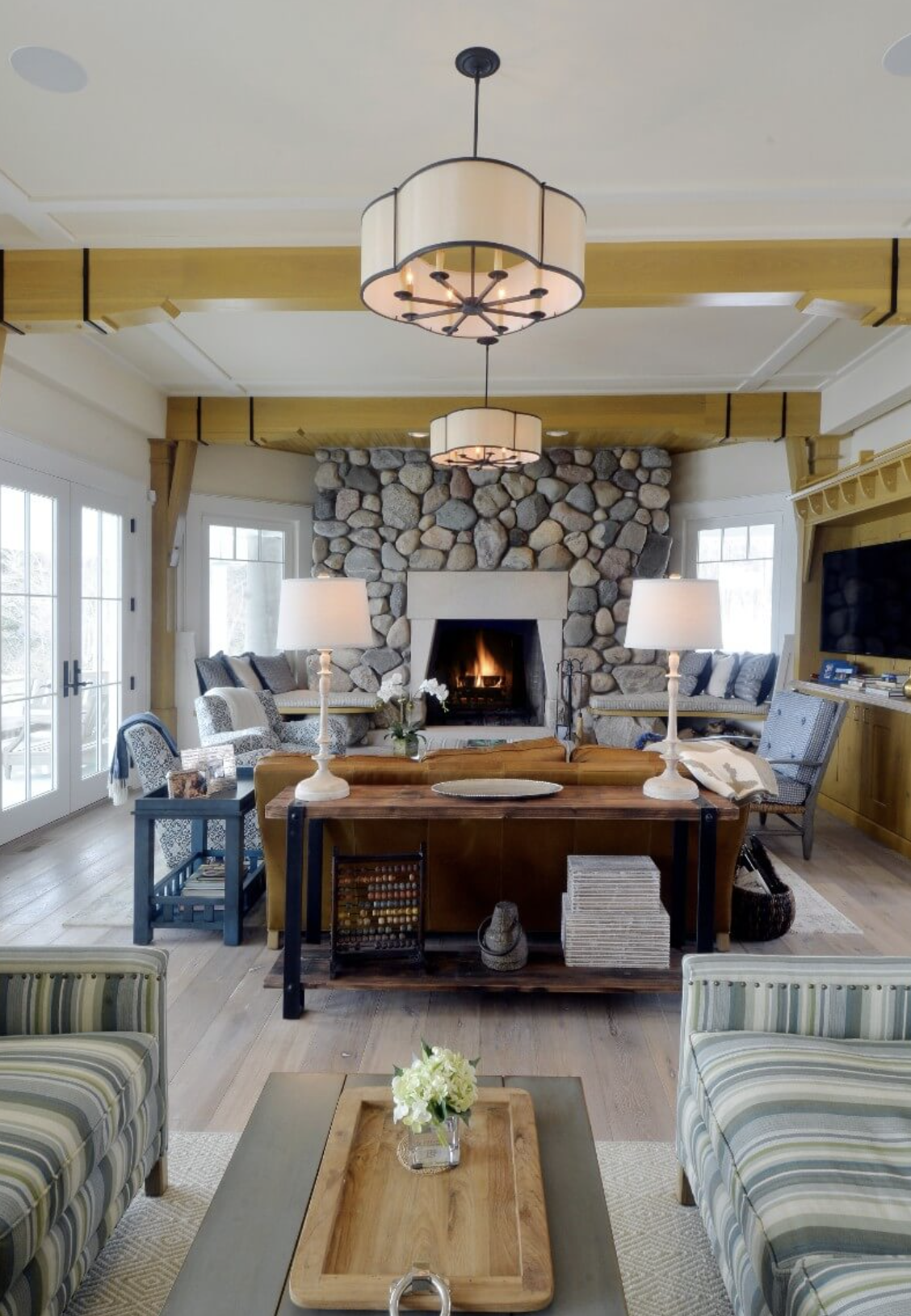 Living room with stone fireplace, arranged sofas, wooden tables, lamps, and large windows.