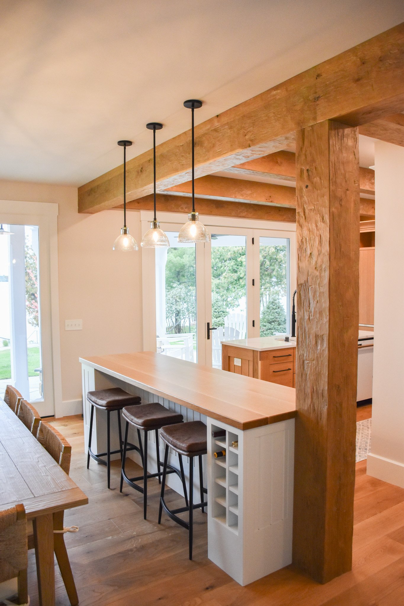 Open kitchen and dining area with large wooden beams, white cabinets, a breakfast bar with four bar stools, pendant lights, and sliding glass doors leading to a backyard with trees.