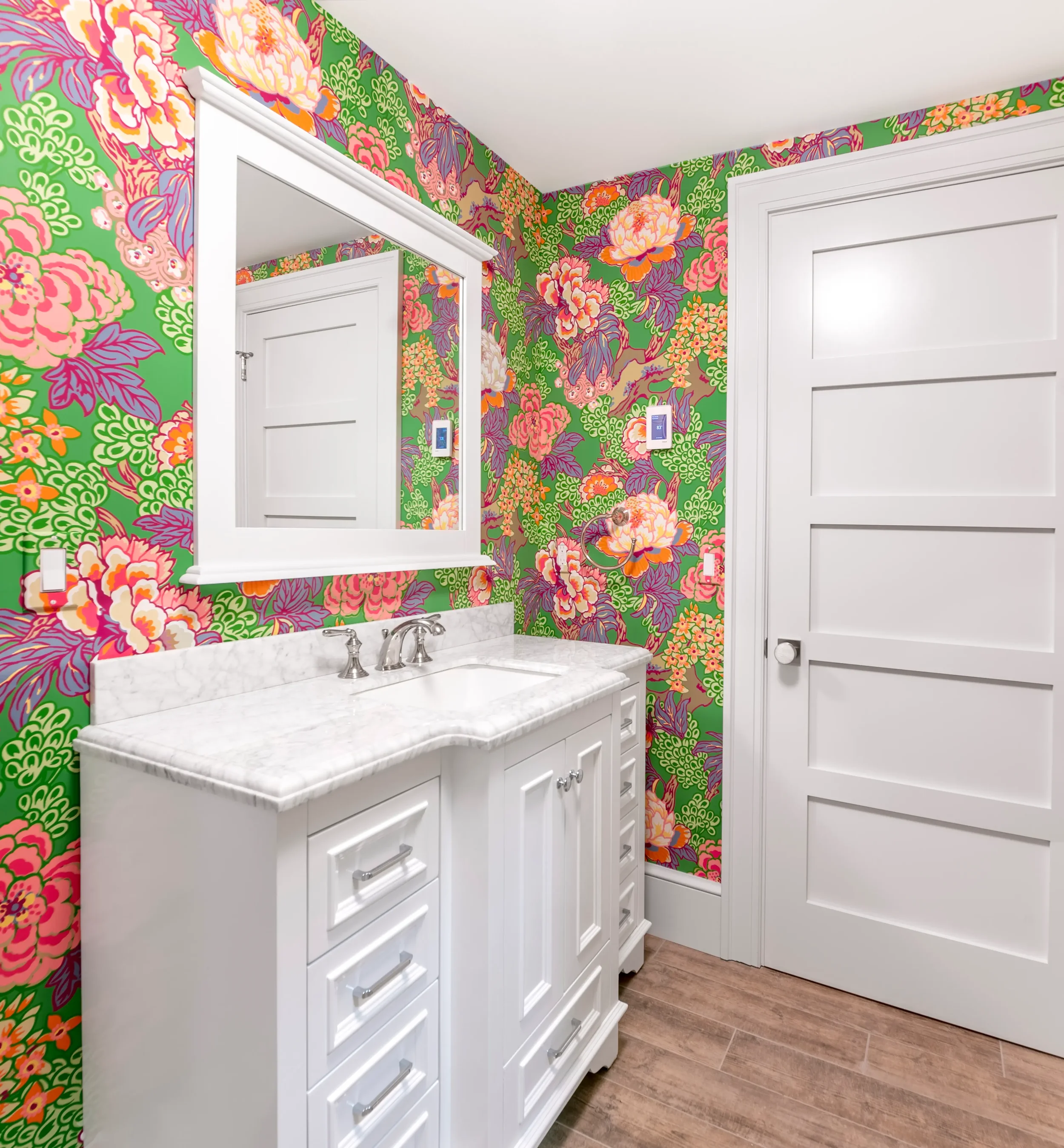 A bathroom with colorful floral wallpaper in pink, orange, purple, and green, a white vanity with a marble countertop and silver hardware, a mirror, and a white door.