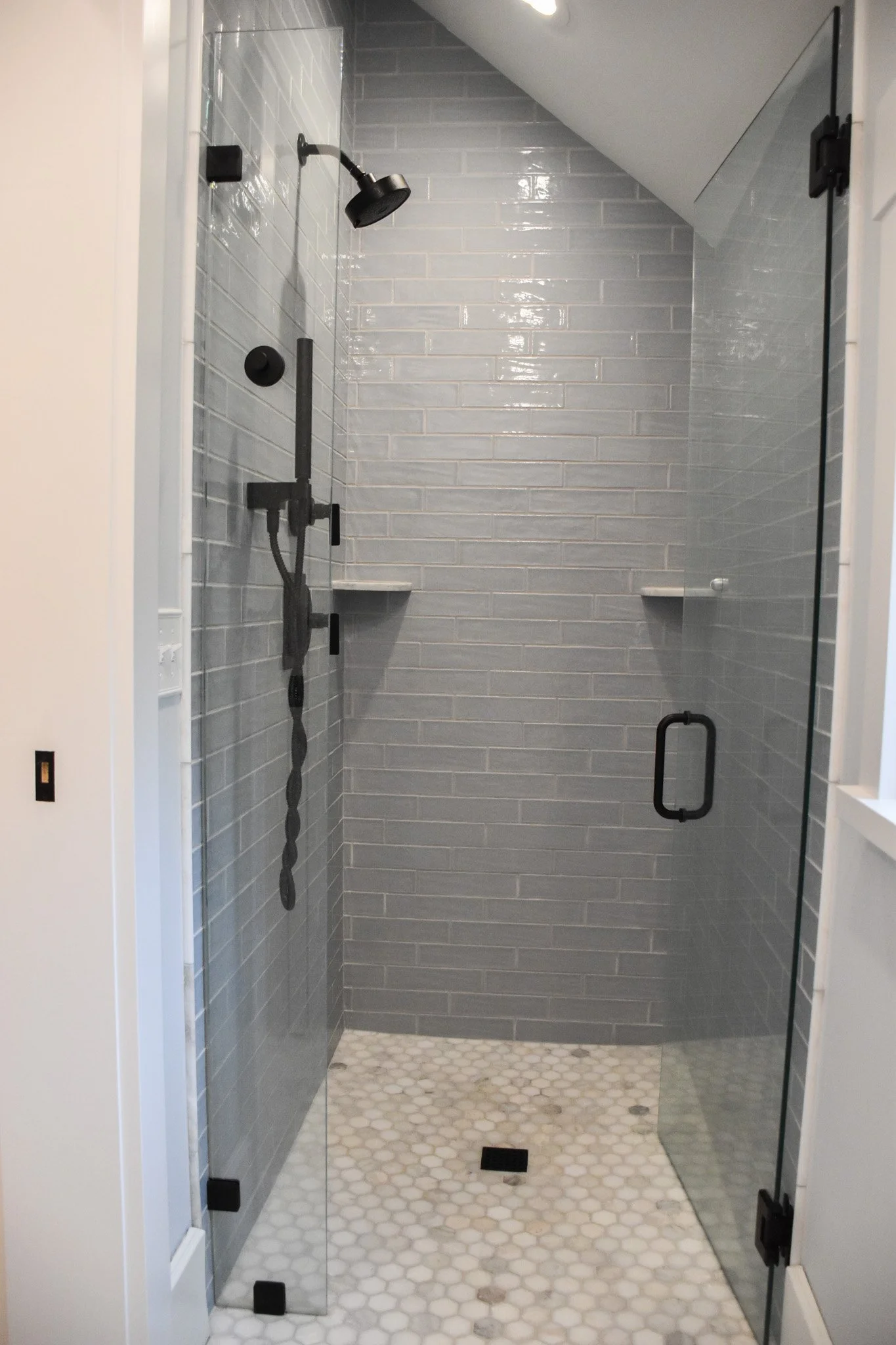 A modern shower with gray tiles on the walls, a black showerhead, and a glass door. The floor has white hexagonal tiles, and there are black accents such as hinges and a handle.