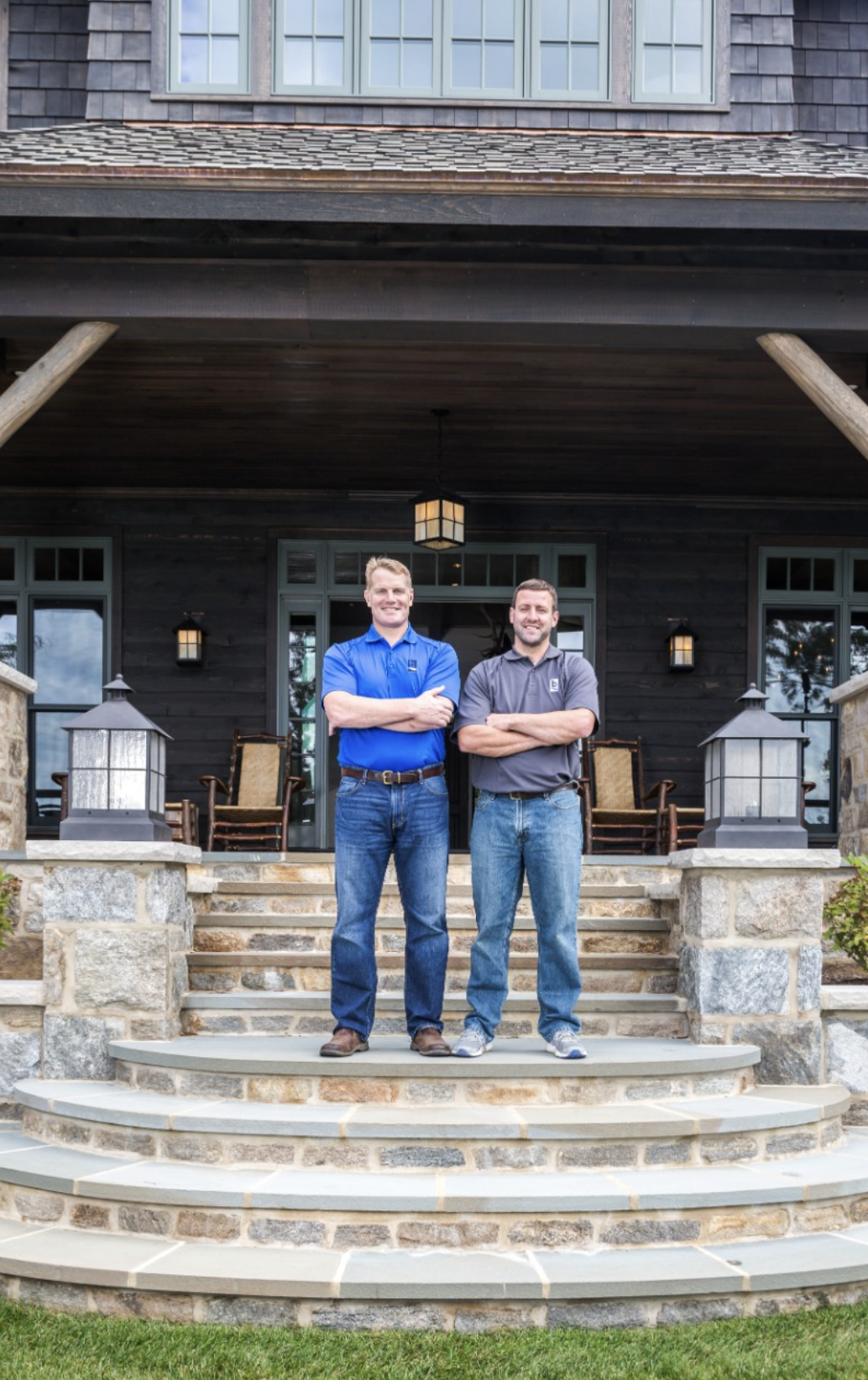 Two men stand on the steps of a porch in front of a black house with large windows, black light fixtures, and outdoor furniture.