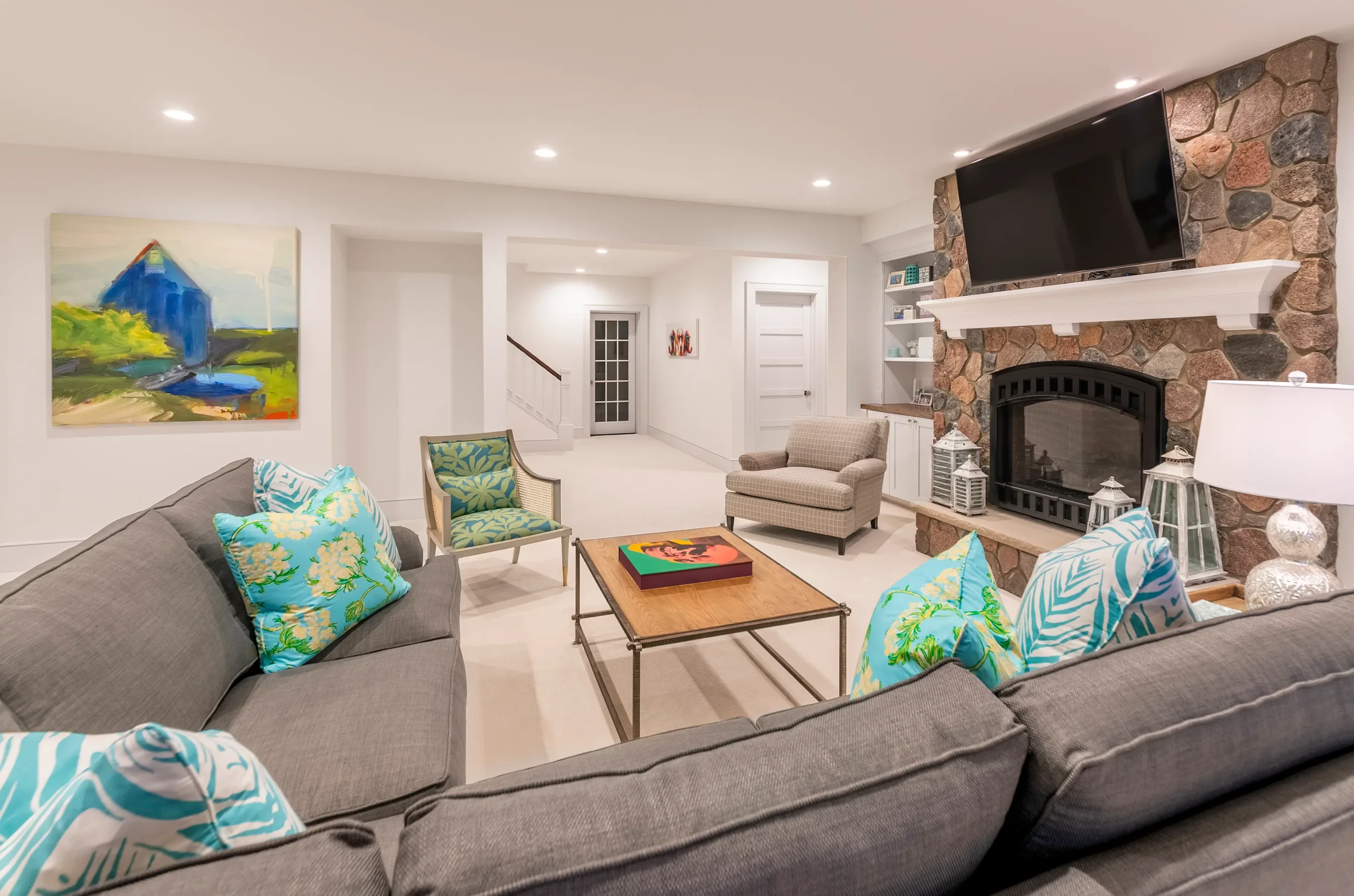 Living room with a gray sectional sofa with colorful blue and green pillows, a wooden coffee table, an armchair, a stone fireplace with a mounted flat-screen TV, a white bookshelf, and artwork on the walls.