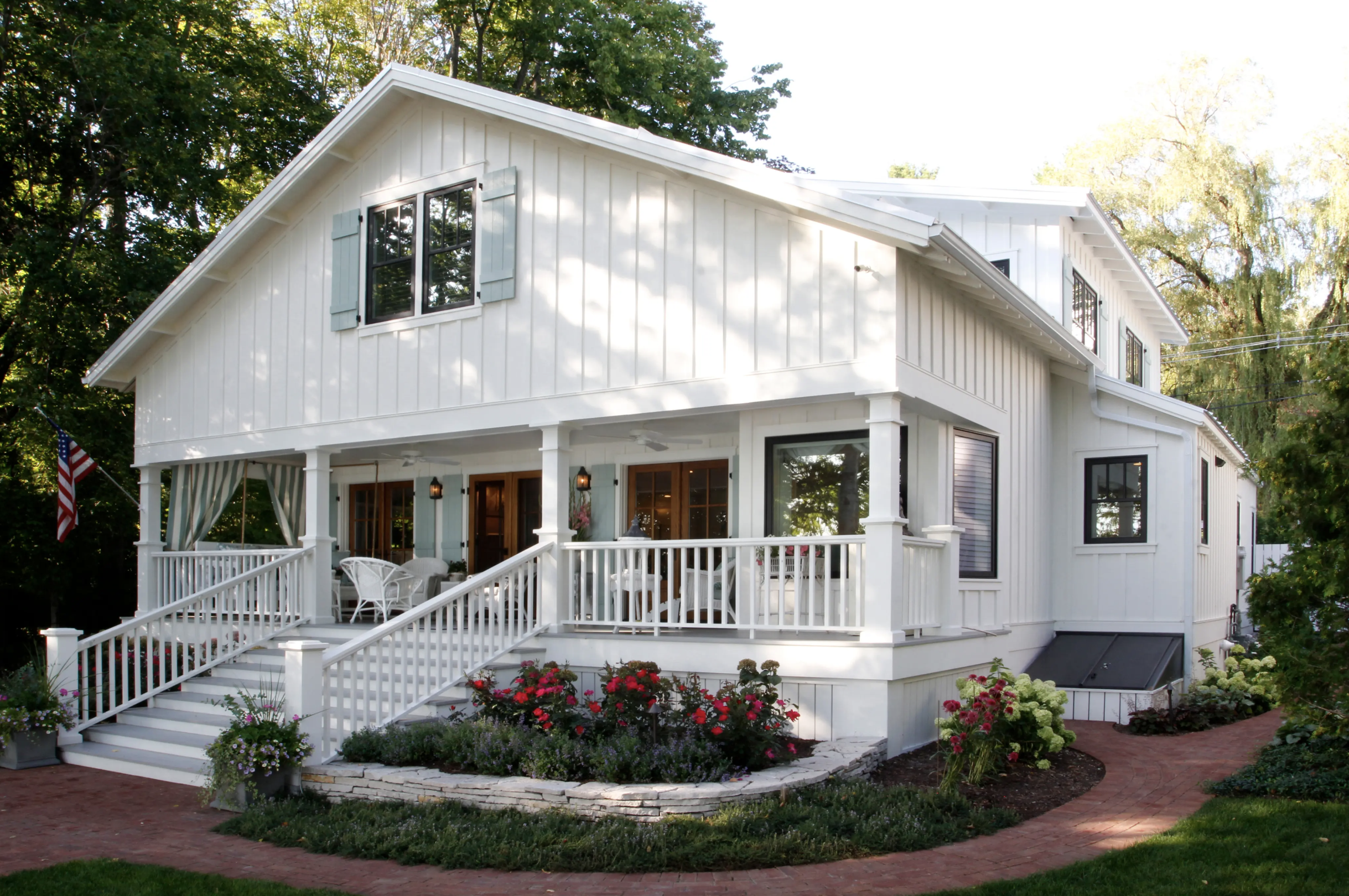 A white two-story house with a porch and a set of stairs leading to the entrance. The house has black window frames, light blue shutters, and a garden with colorful flowers and bushes. An American flag is visible to the side.