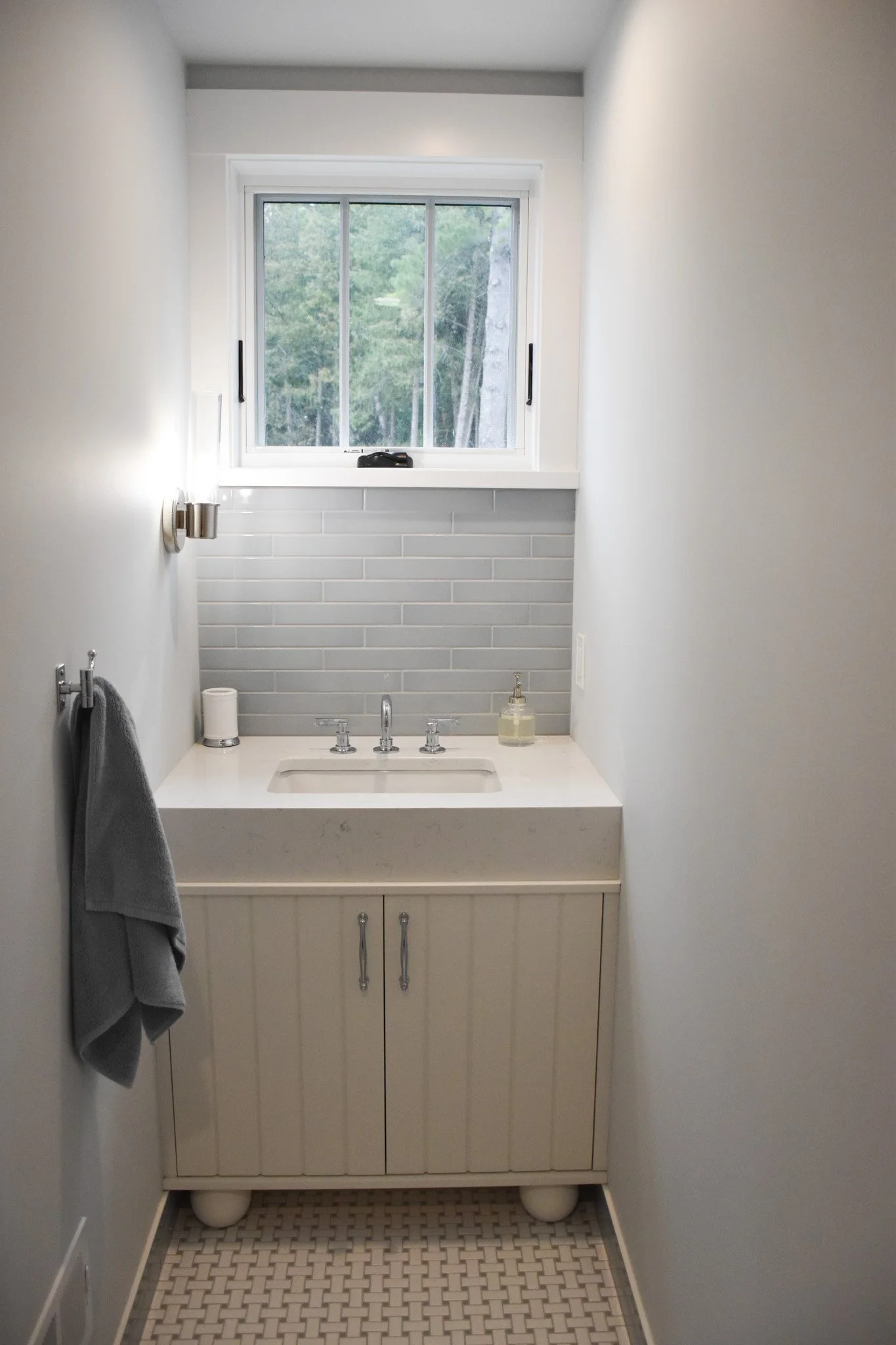 Small bathroom with a window above a white vanity with a backsplash of white subway tiles. There are soap and hand sanitizer bottles on the sink. Grey walls with a towel hanging on a hook on the left side.