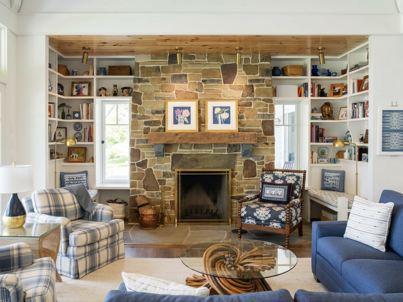 Living room with a stone fireplace, surrounded by white bookshelves filled with books and decorative items, decorated with framed floral art above the mantle, and furnished with patterned sofas and armchairs, a glass coffee table, and brass lamps.