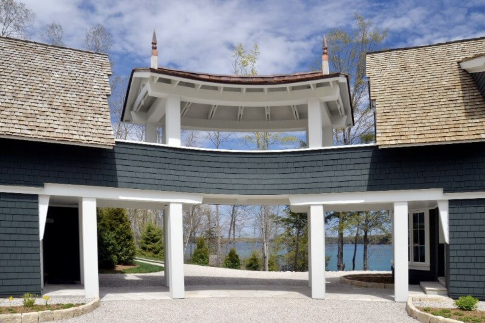 View of a modern house with a central open-air pavilion and water in the background, showcasing a balcony, columns, and roof details.