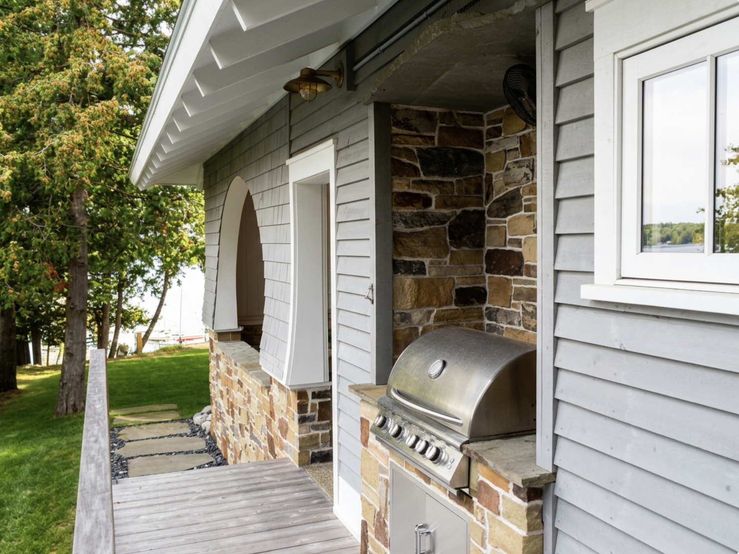 Part of outdoor patio area with a stainless steel grill, stone accents, a white window, and a wall light fixture, with trees and grass in the background.