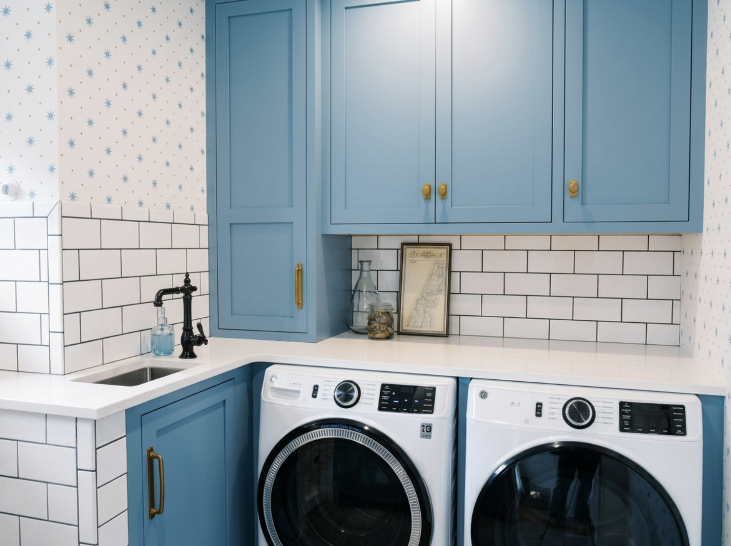 A laundry room with blue cabinetry, a white countertop, white subway tile backsplash, a small sink with a black faucet, and a front-loading washing machine and dryer. There are decorative jars and framed artwork on the counter.