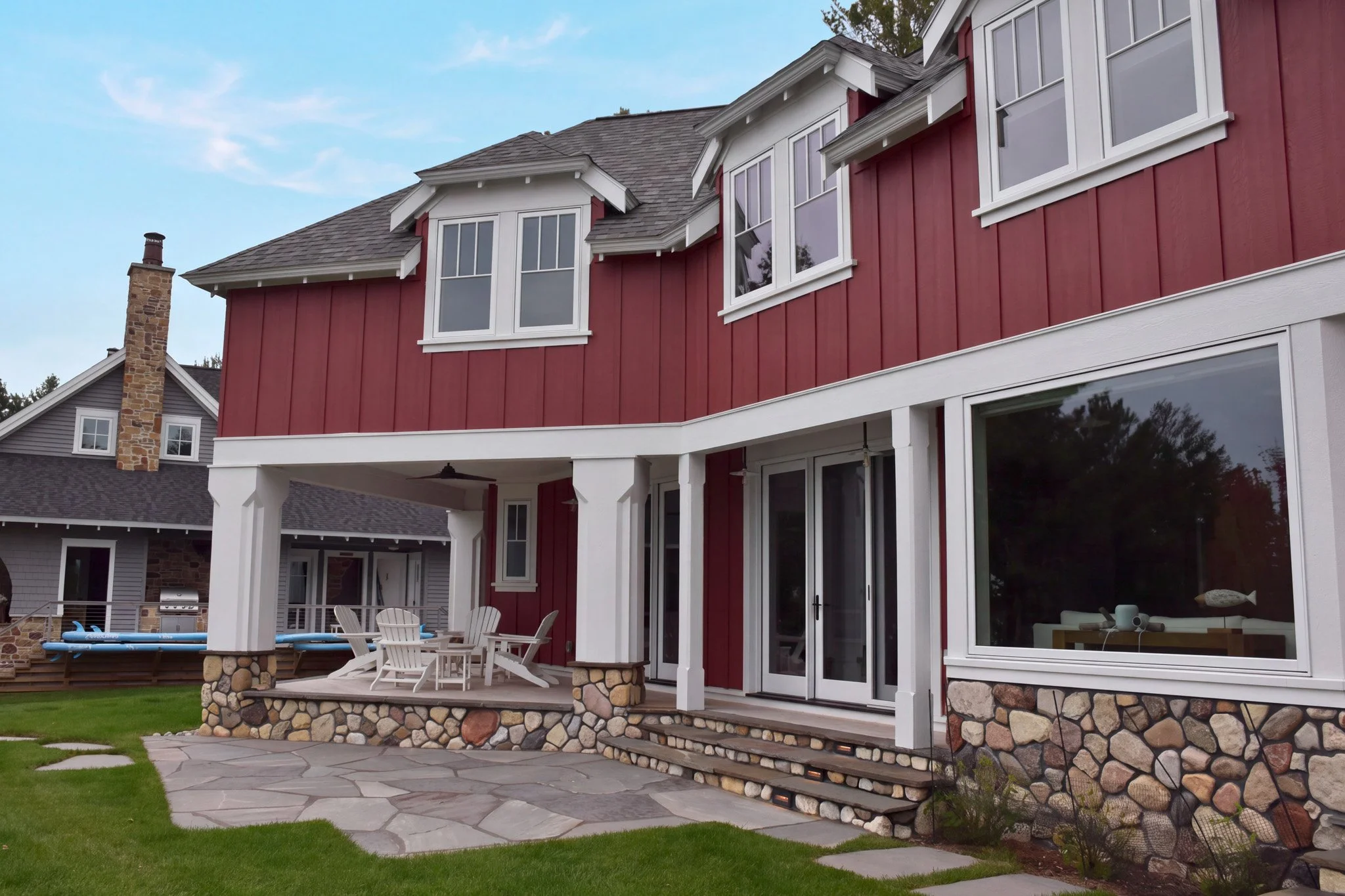 A backyard patio of a house with red siding, white trim, and large windows. There is outdoor furniture, a stone fire pit, a stone staircase, and a lawn with a stone pathway.