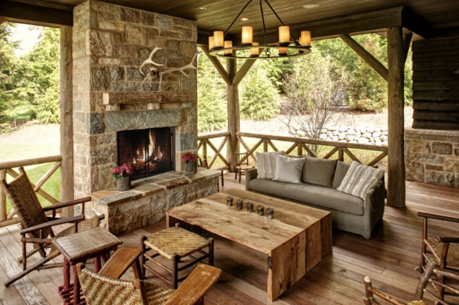 A cozy outdoor porch with a stone fireplace, wooden furniture, and a ceiling chandelier surrounded by trees.