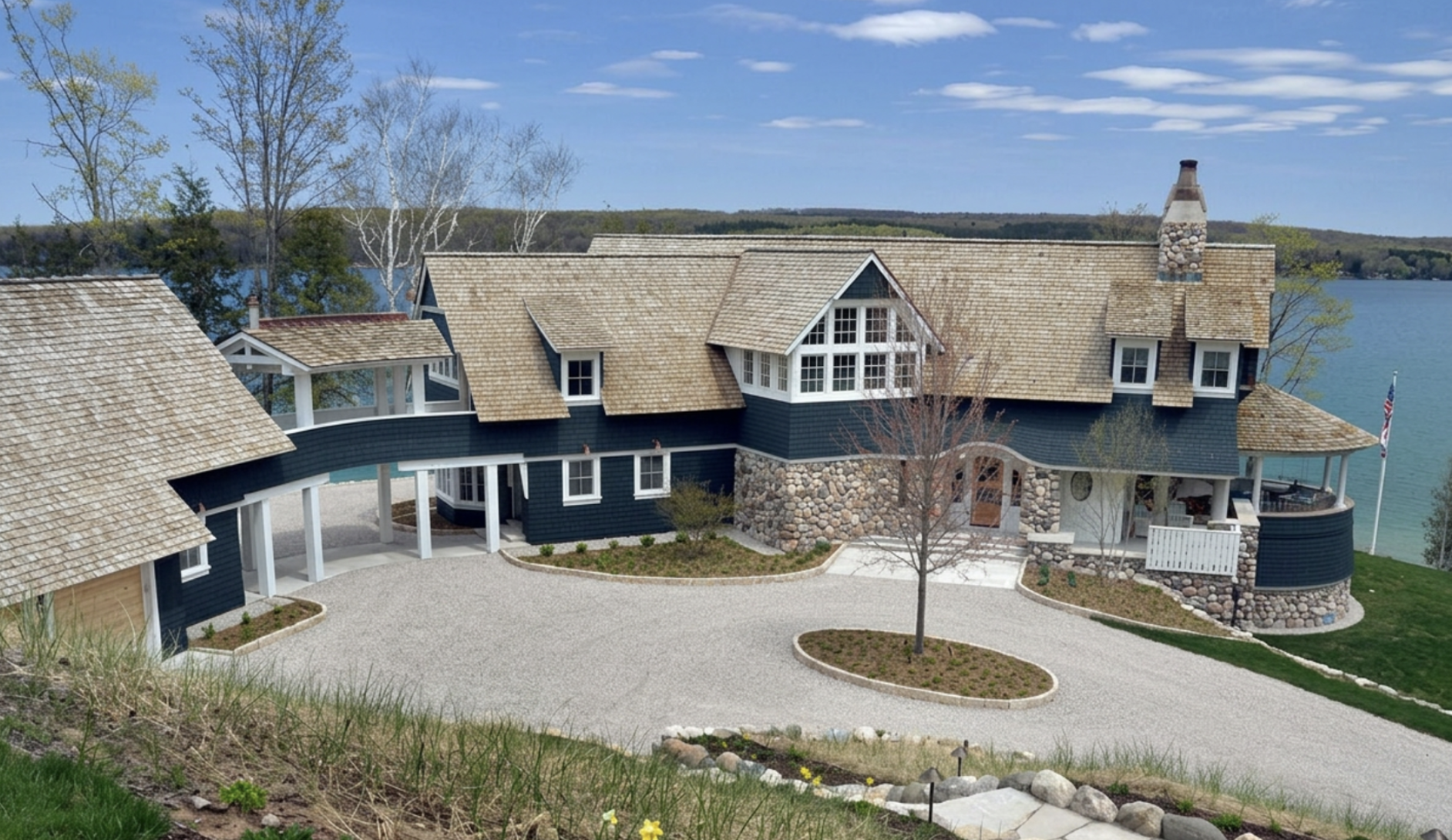 A large lakeside house with a stone foundation, dark blue siding, and multiple gabled roofs with tan shingles, surrounded by a gravel driveway, landscaped garden beds, and trees, with a view of water and hills in the background.