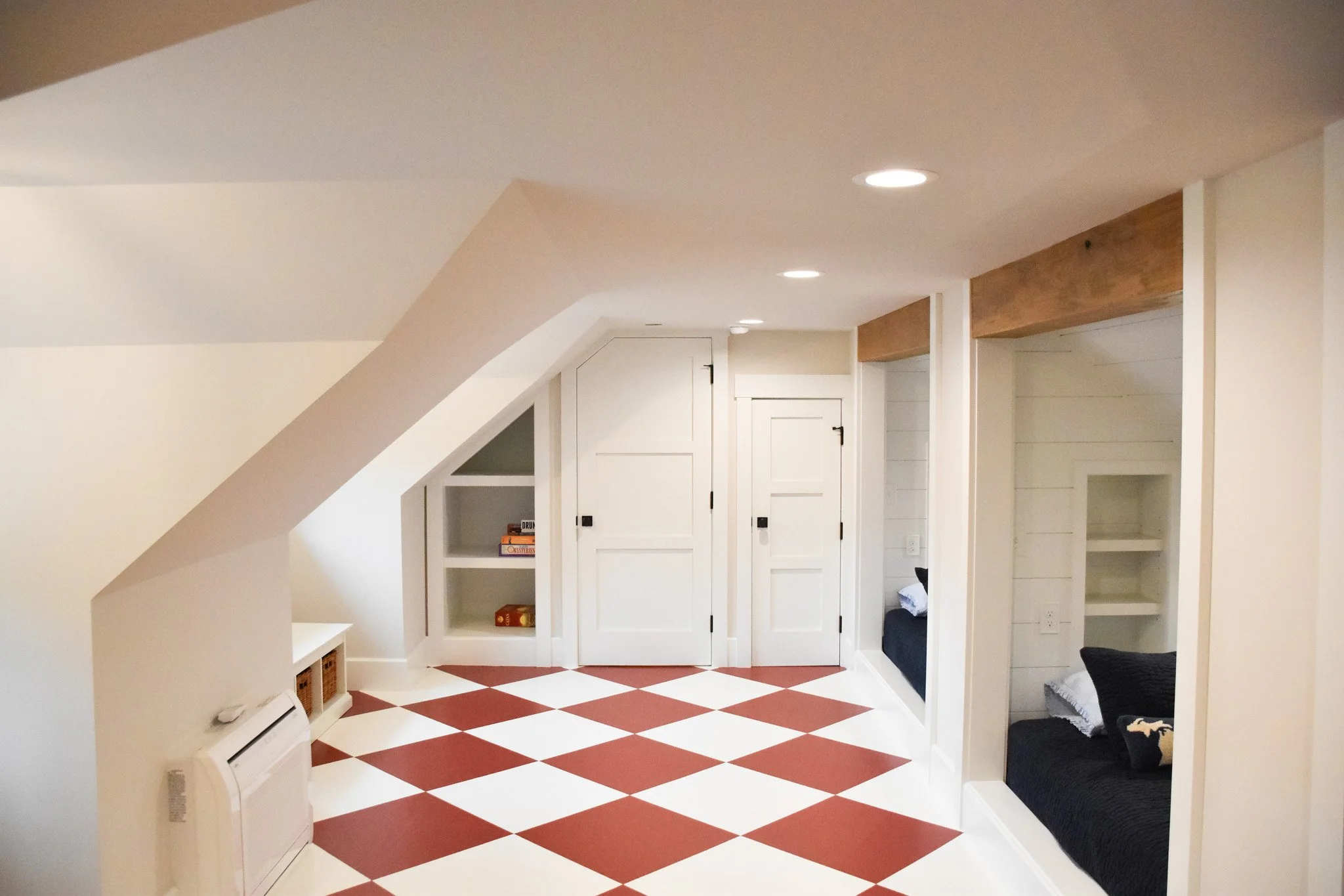 A cozy attic living space with white walls and sloped ceiling, featuring white built-in cabinets, a black bench with a white blanket, and a red and white checkered tile floor.
