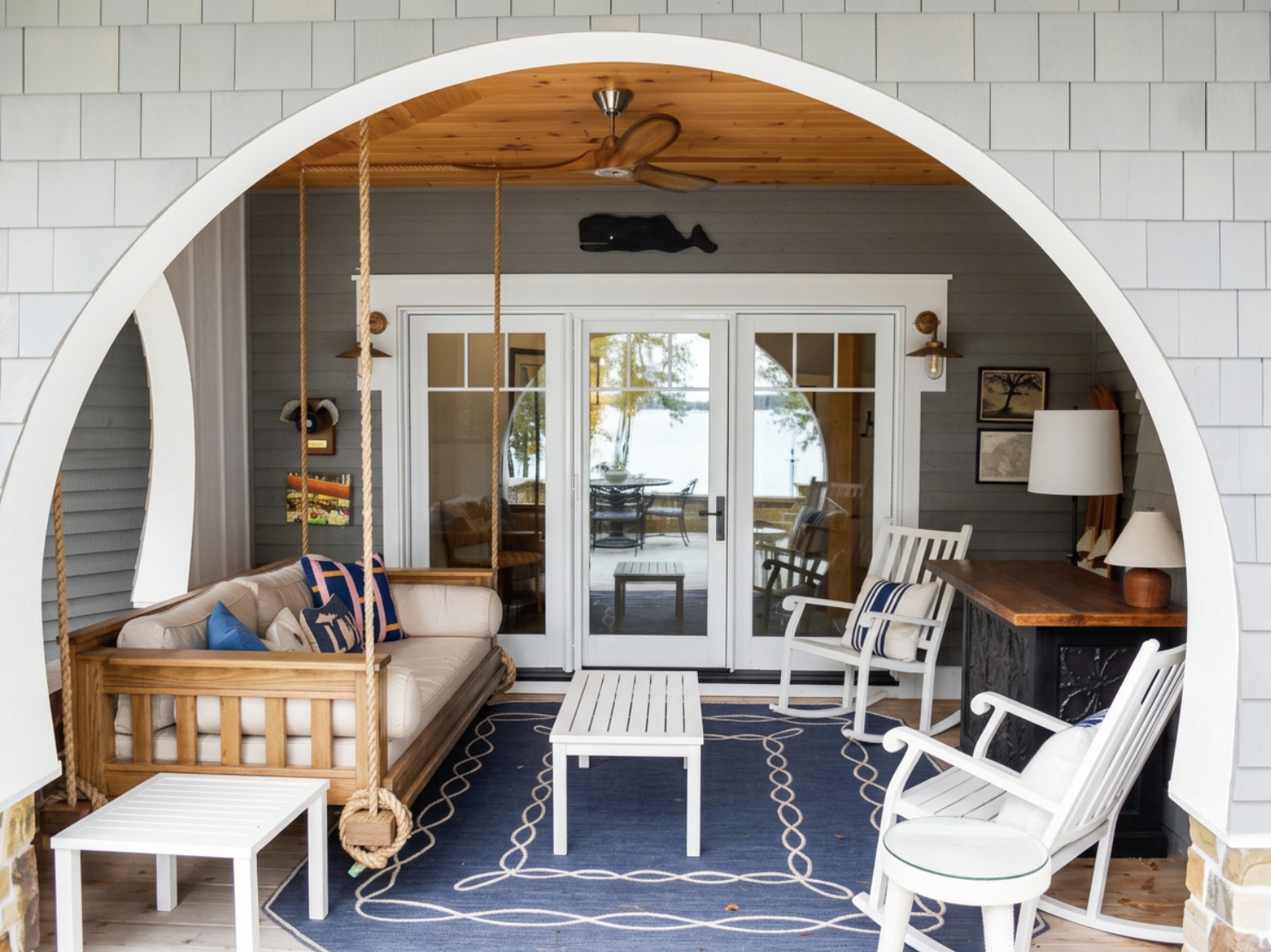 Covered porch with hanging swings, white chairs, wooden table, and view of a lake through glass doors.