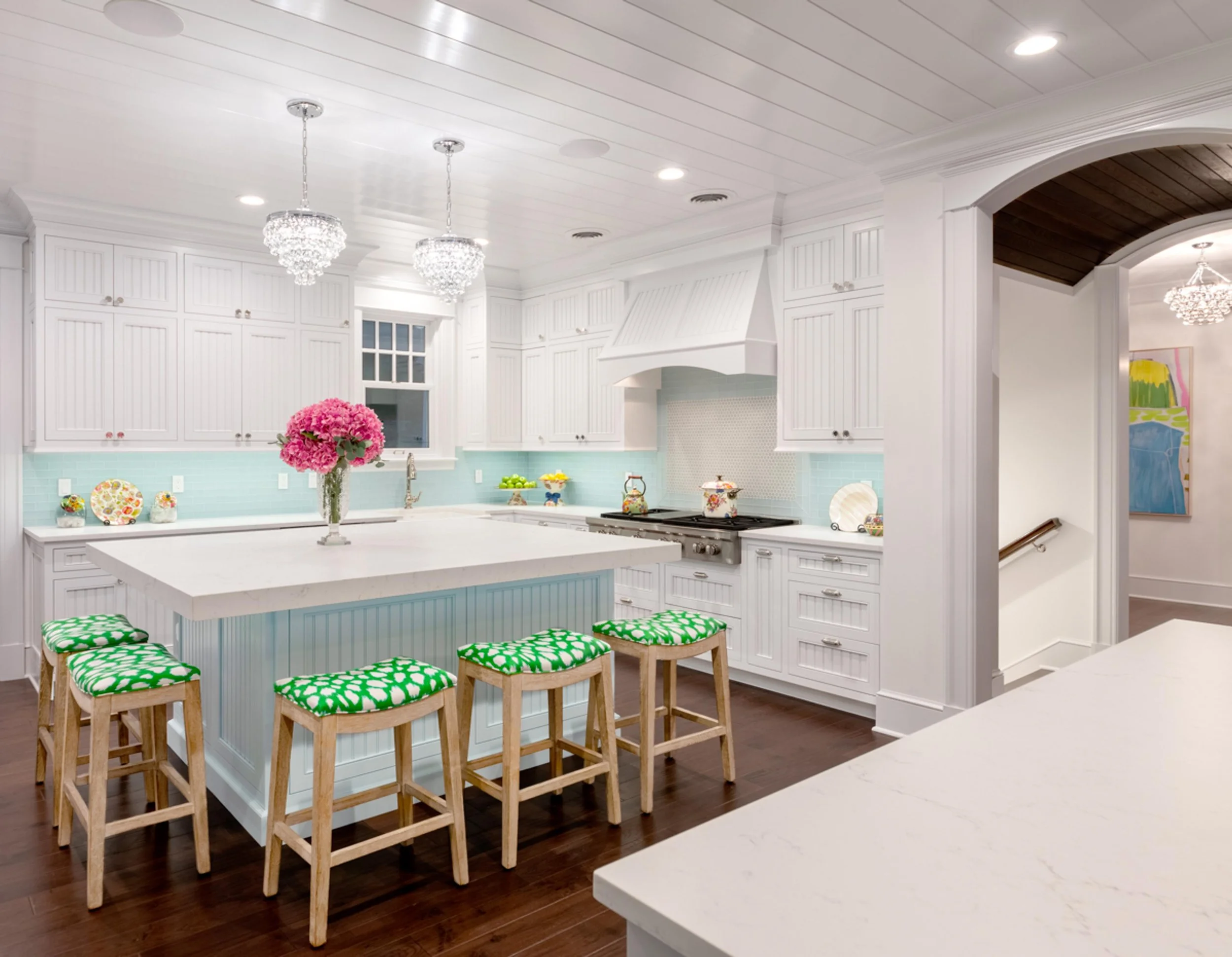 Modern kitchen with white cabinetry, a white island with green and white patterned stools, and light blue subway tile backsplash. Two crystal chandeliers hang from the ceiling, with a bouquet of pink flowers on the island. There is a doorway with a w