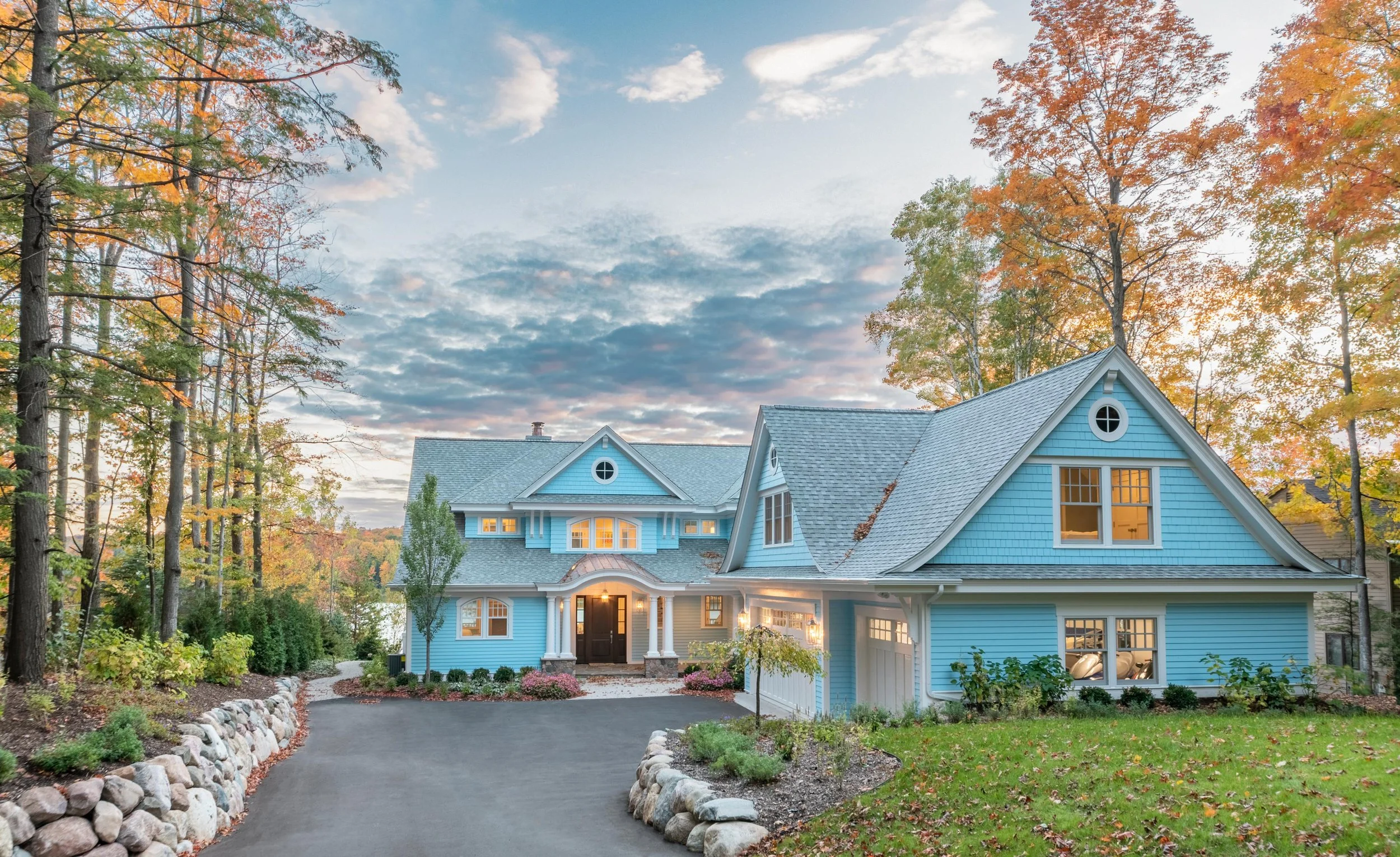 A large blue house with a steep grey roof and white trim, surrounded by trees with autumn leaves, and a curved driveway lined with rocks leading to the front entrance, illuminated by warm outdoor lighting.