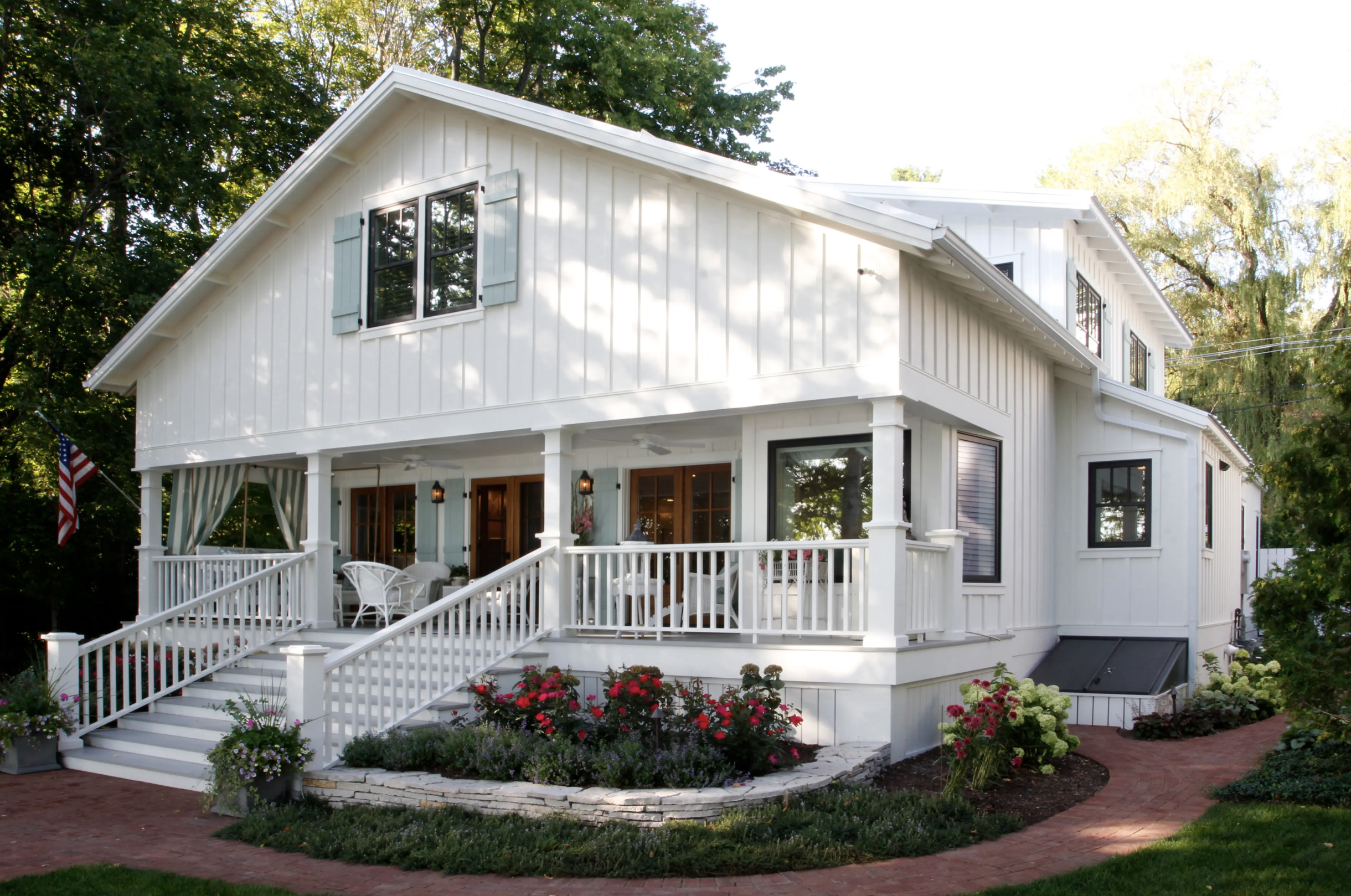 A two-story white house with a porch, large windows, and shutters, surrounded by a garden with colorful flowers, and trees in the background.