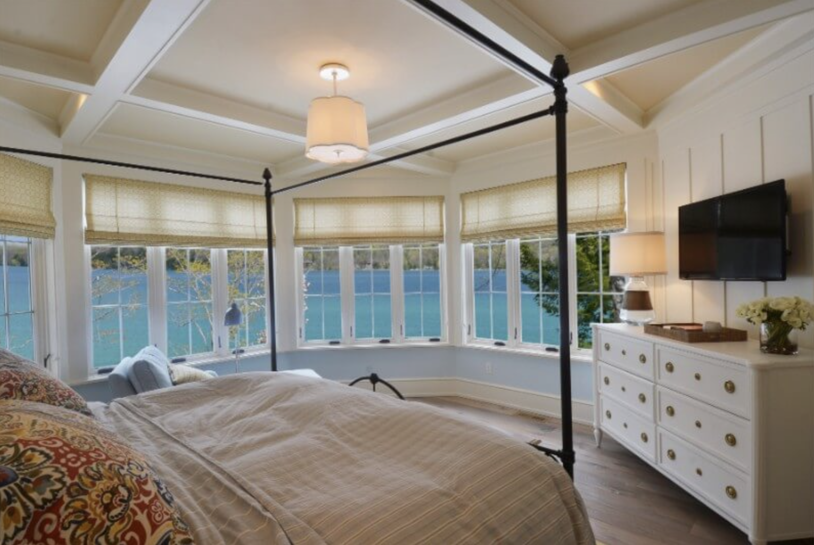 A cozy bedroom with a large bed, a white dresser, a flat-screen TV, and large windows showing a view of water and trees outside.