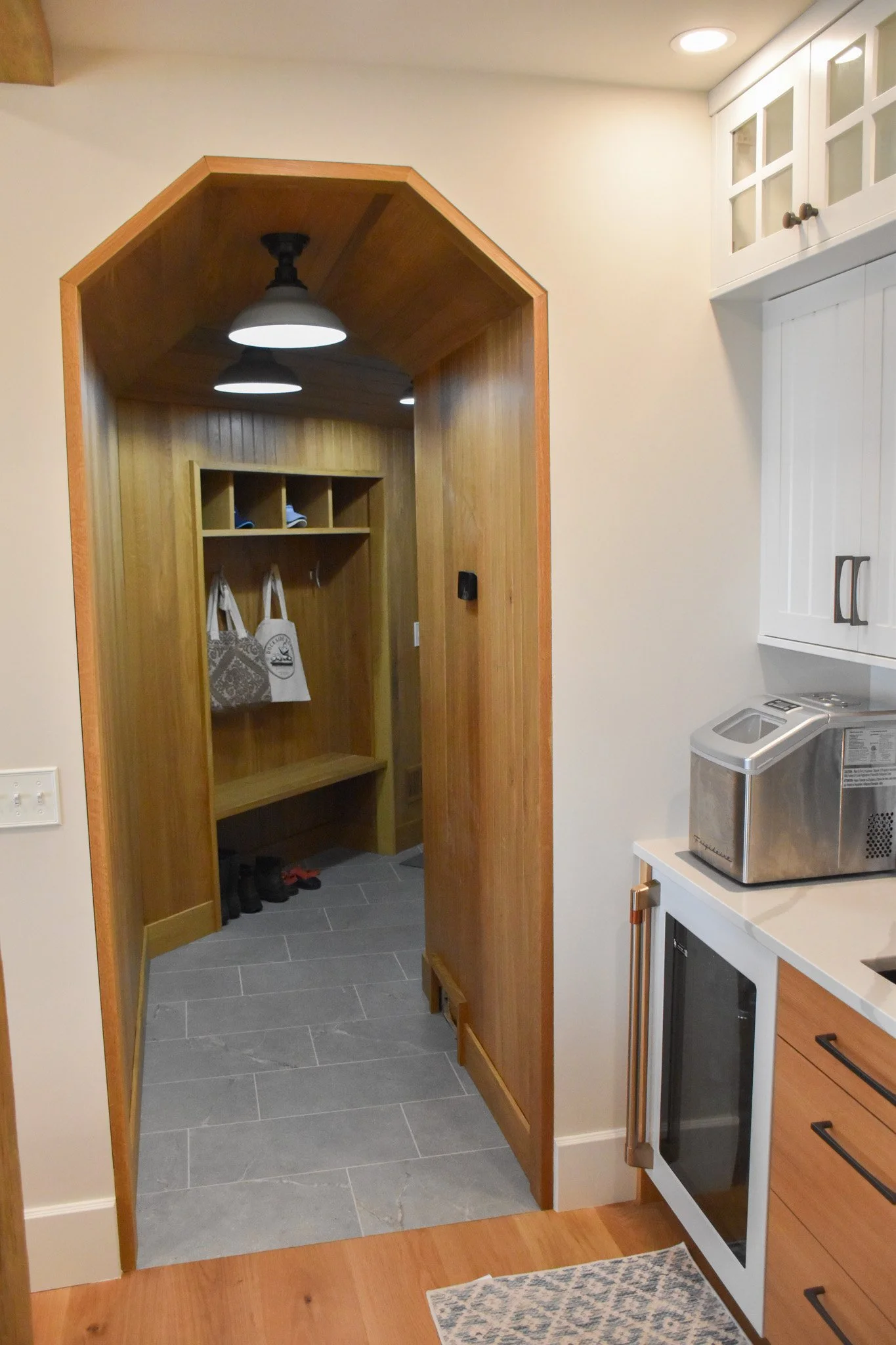 A kitchen doorway with a wooden frame leads into a small mudroom with wood-paneled walls and a shelf with hooks holding shoes and bags. The room has a gray tile floor and hanging ceiling lights.