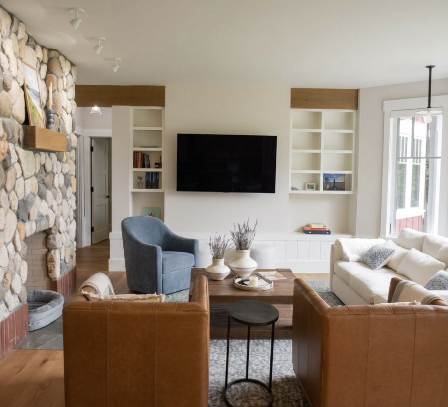 Living room with stone fireplace, white built-in shelves, mounted flat-screen TV, beige sofa with pillows, blue armchair, wooden coffee table with vases and books, window with view outside.