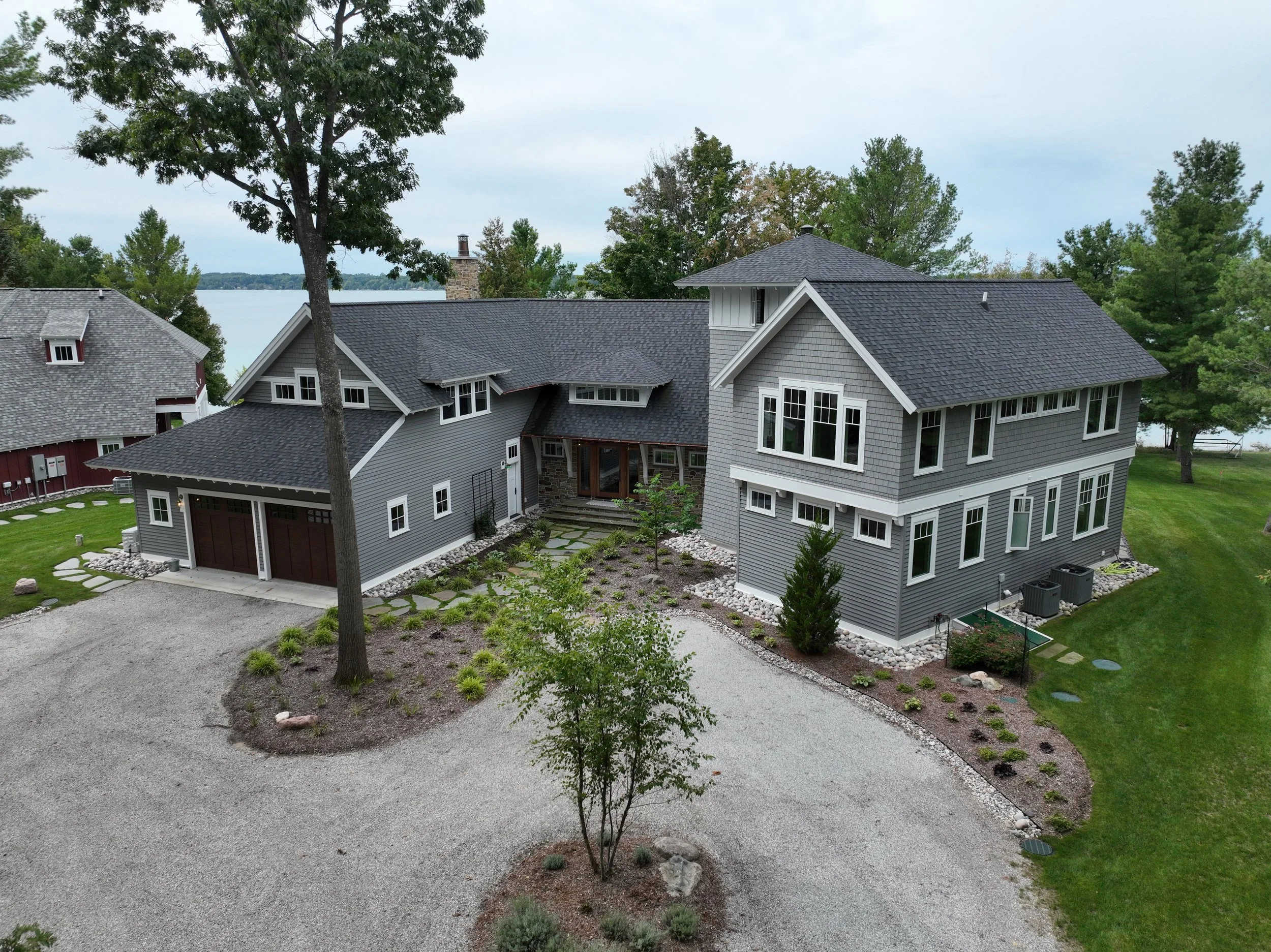 A two-story gray house with a dark gray roof, multiple windows, and a stone chimney, adjacent to a red barn, set in a landscaped yard with trees, a gravel driveway, and a view of water in the background.