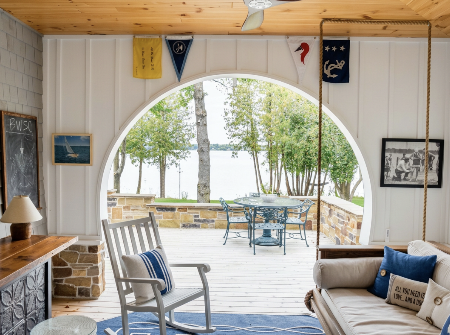 A cozy indoor space with a view of an outdoor patio near a body of water. The room features a wooden ceiling, white paneled walls, and nautical-themed decorations, including flags and framed pictures. There is a white rocking chair with a striped pil