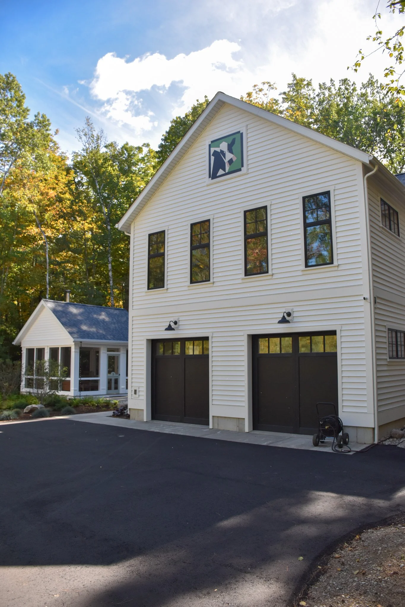 White house with black garage doors and a sign of two people playing a game, overlooking trees with green and orange leaves on a sunny day.