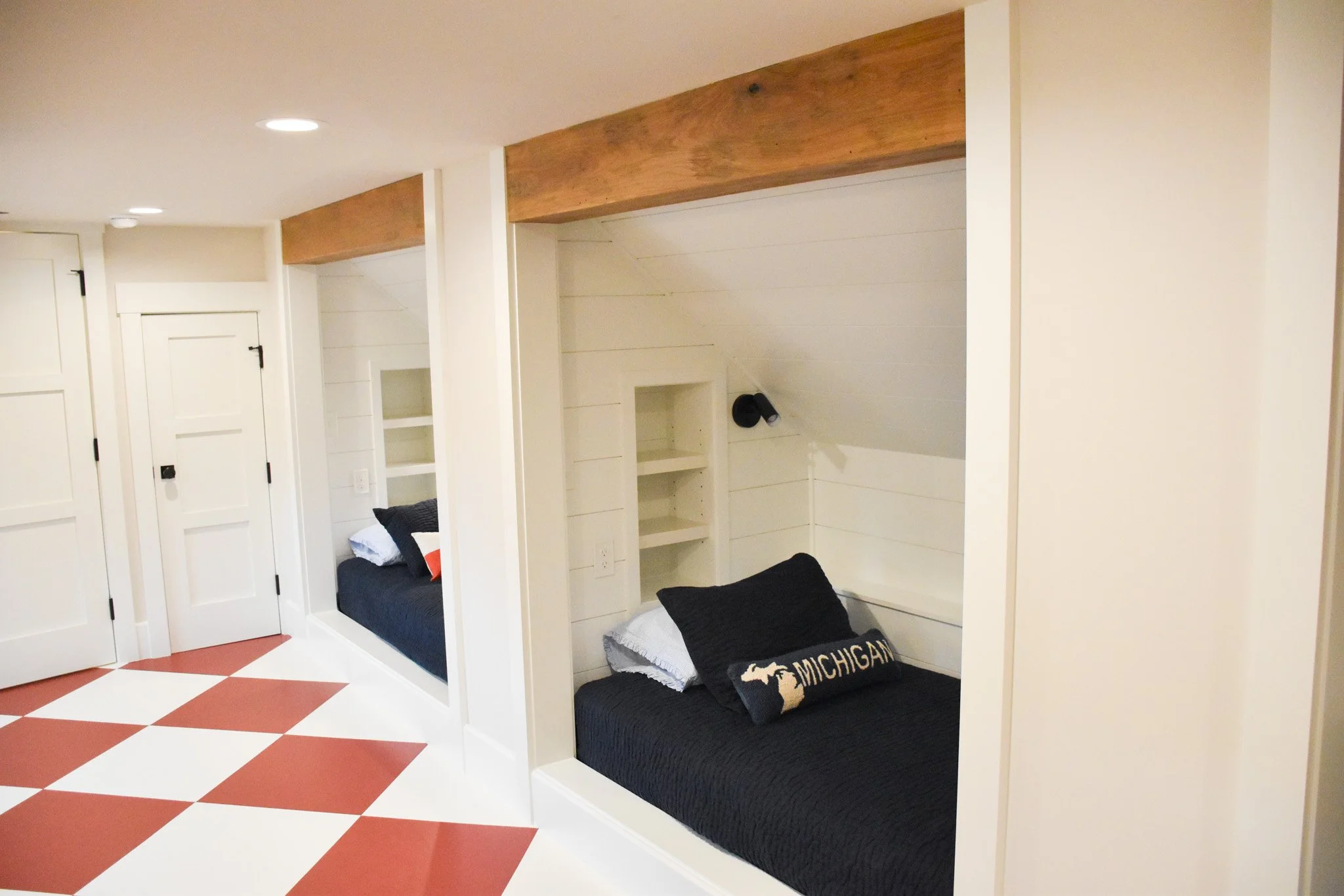 Interior of a room with built-in beds within alcoves, white walls, and a wooden beam ceiling. The beds have black mattresses and pillows, and one pillow features a Michigan state design. The room has a checkered red and white tile floor.