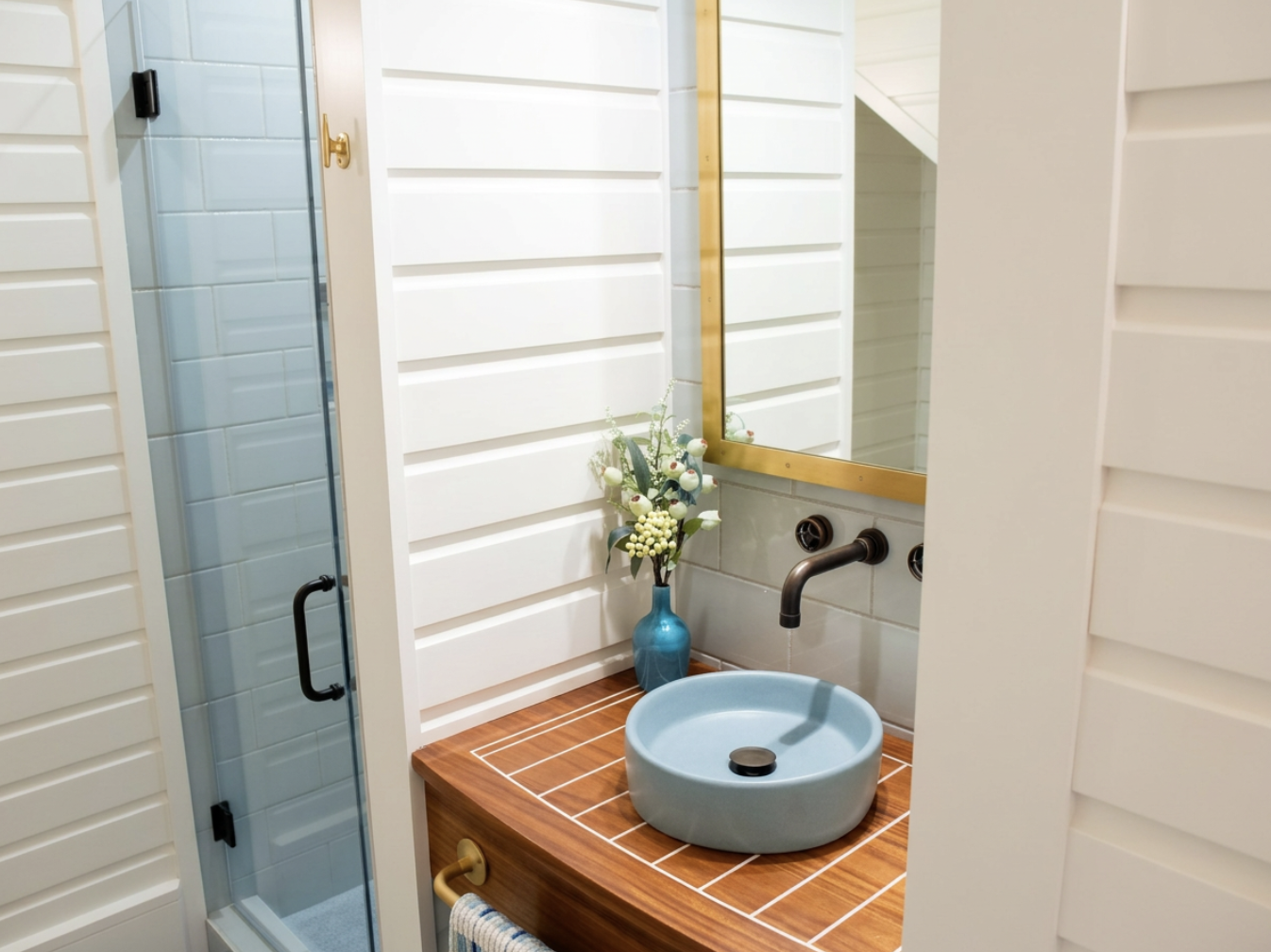 A bathroom vanity with a round blue vessel sink, a bronze faucet, a mirror with a gold frame, a blue vase with white and green flowers, and wooden countertop tiles, with a glass shower on the left.