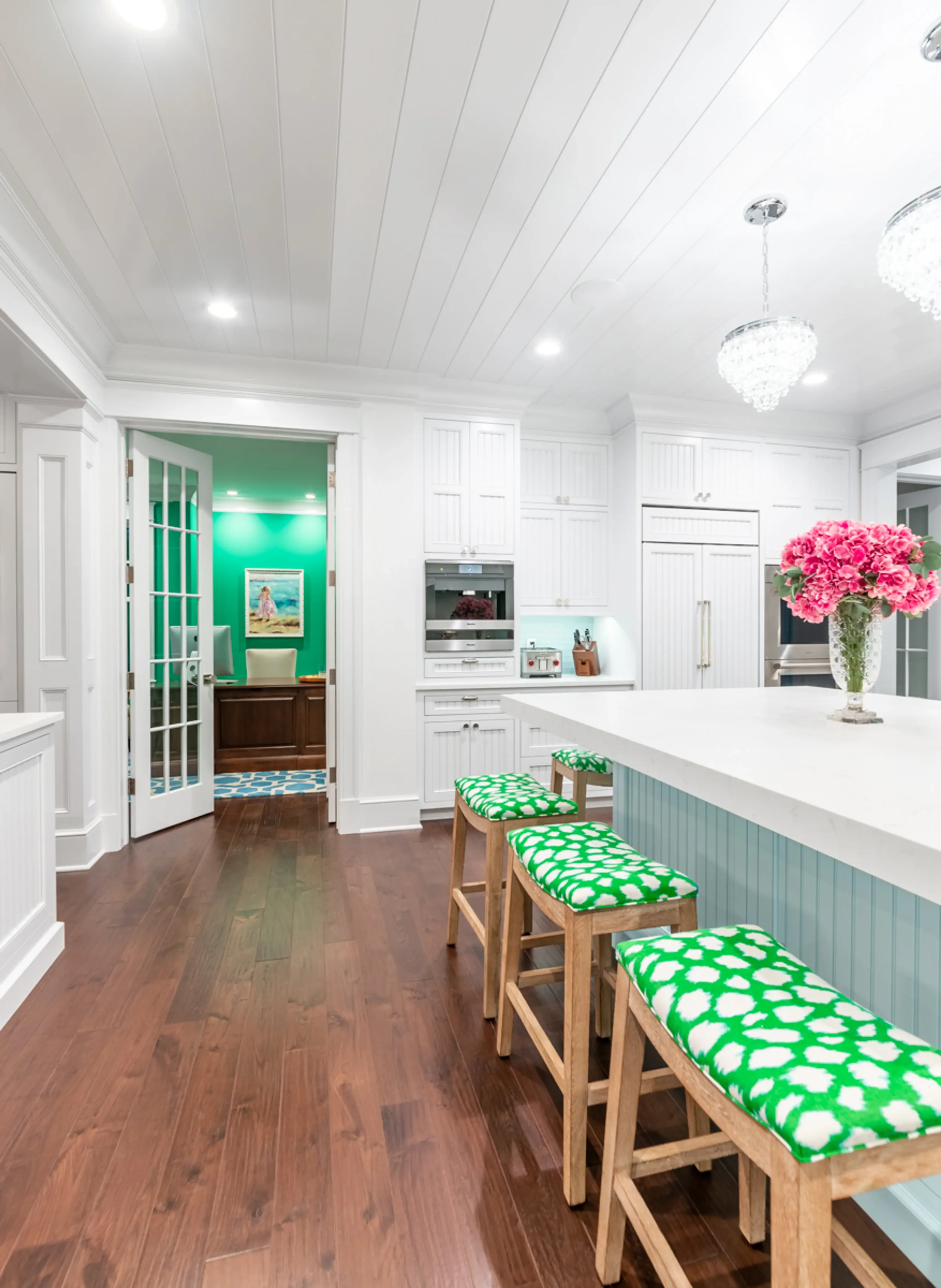 A kitchen with white cabinets, a white kitchen island with a vase of pink flowers, wooden stools with green and white patterned cushions, and a view into a room with green walls and a painting, with hardwood floors and ceiling lights.
