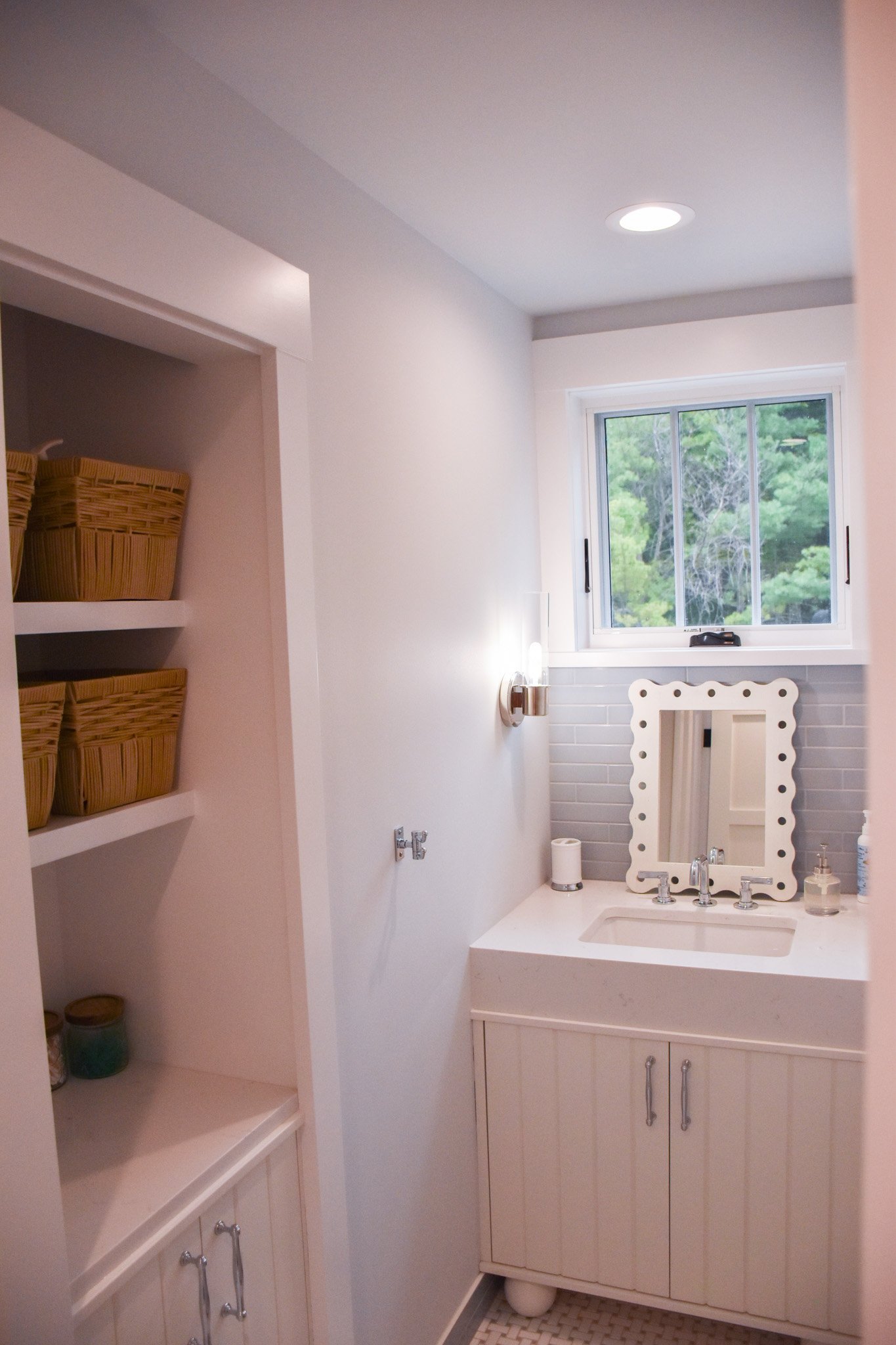 A bathroom vanity with a white countertop, a small mirror with a white frame, a faucet, and a window showing green trees outside. There is a wall lamp, a soap dispenser, and some toiletries on the countertop. To the left, there are built-in shelves w