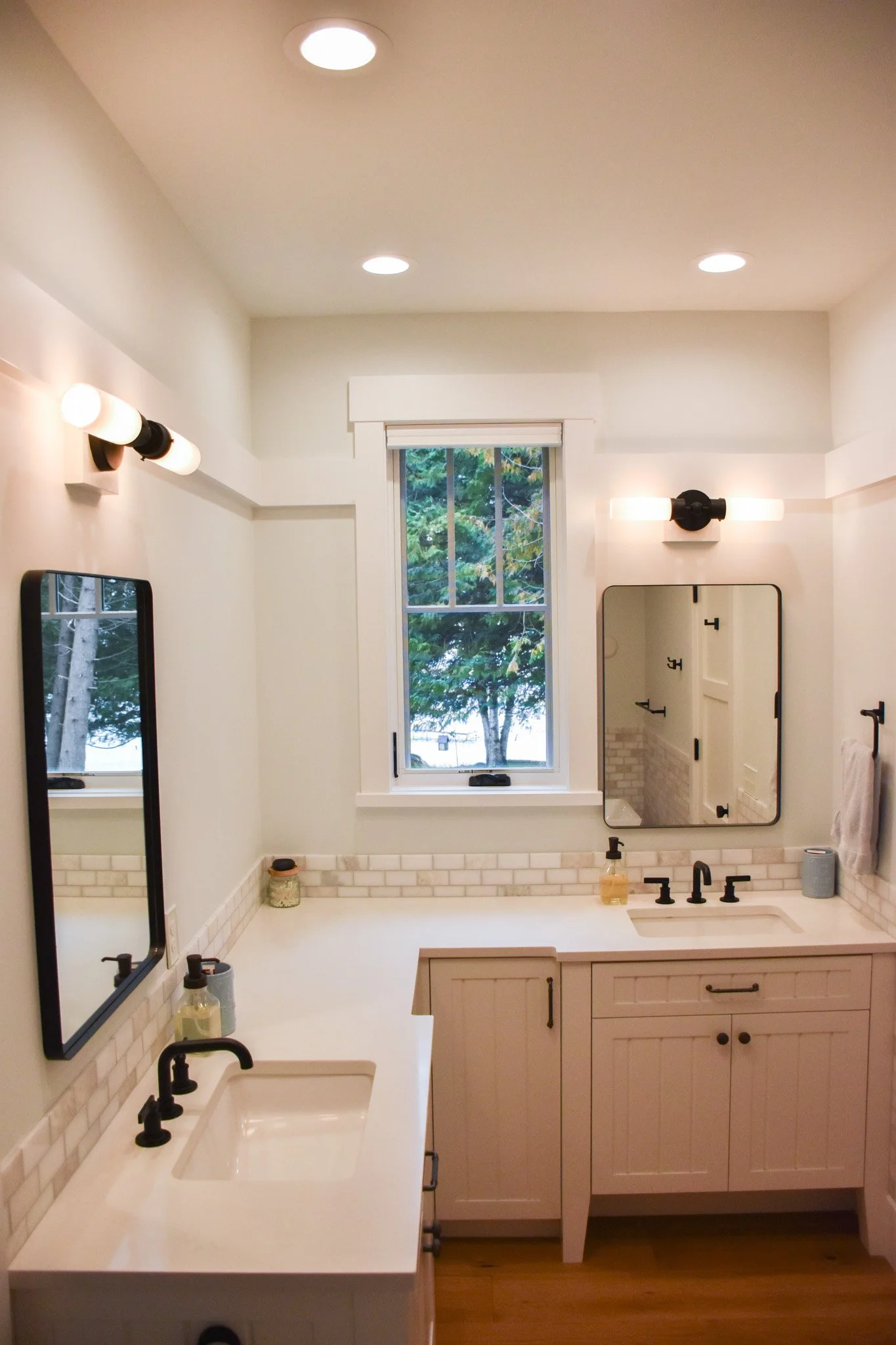 A bathroom with a white vanity, a sink, black fixtures, a large mirror, and a window showing trees outside. The ceiling has three recessed lights and two wall-mounted lights above the mirror. There are soap and lotion bottles on the countertop.