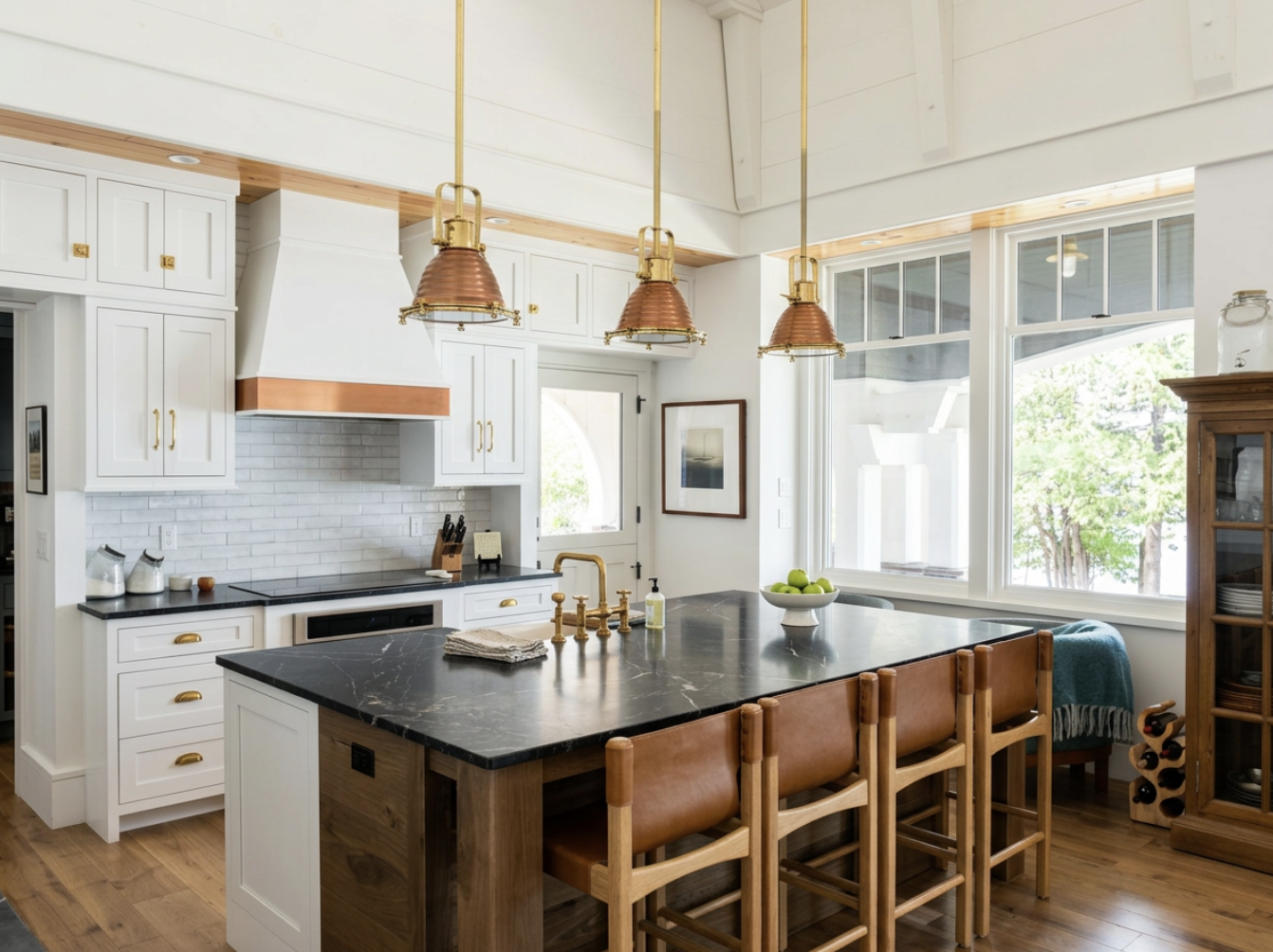 Modern kitchen with white cabinets, black countertop island, brass fixtures, three pendant lights, large windows, wooden chairs, and a wine rack.