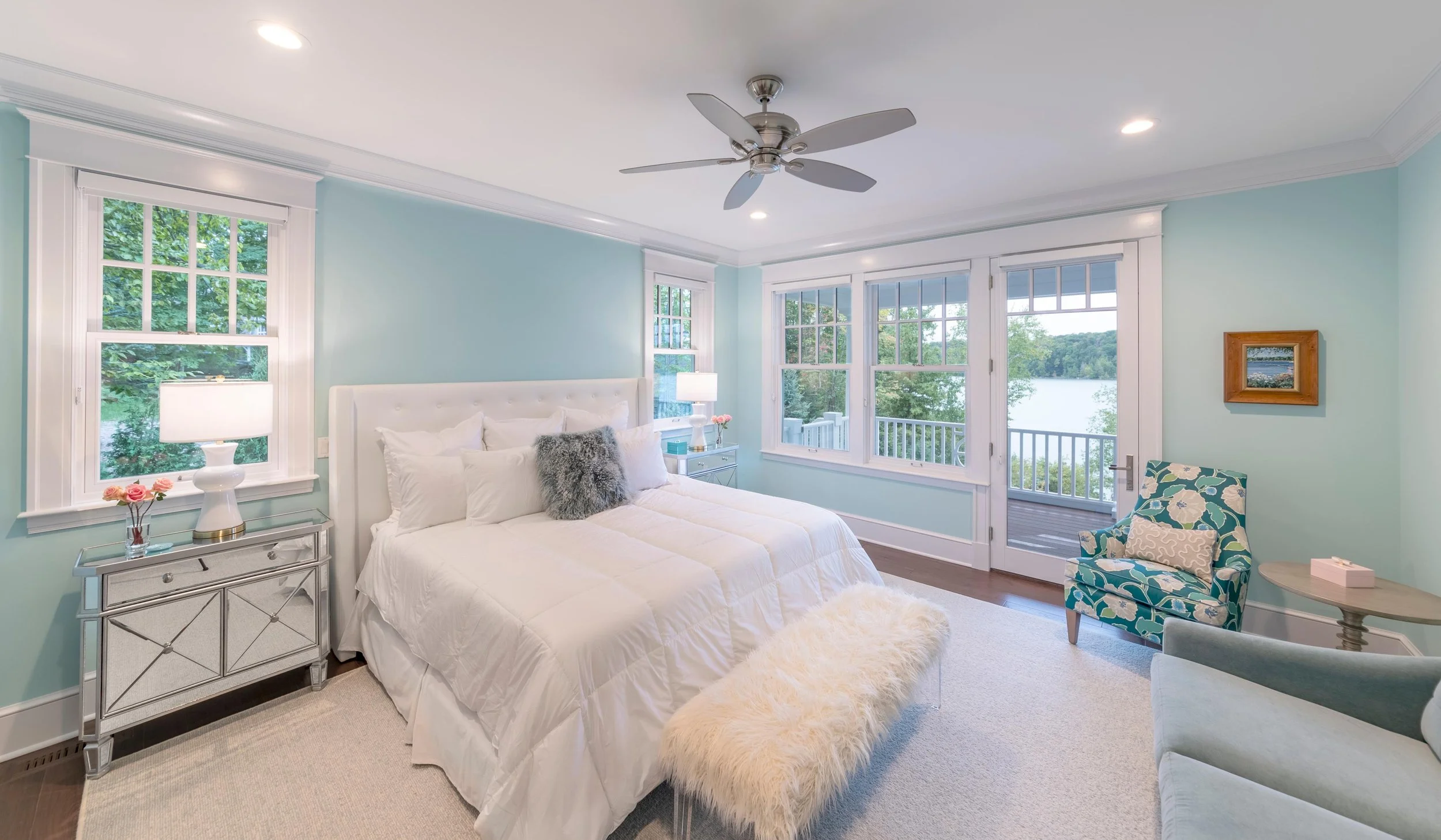 A bedroom with light blue walls, large windows showing a view of water and trees, a white bed with pillows, a mirrored nightstand with a lamp and flowers, a floral armchair, and a ceiling fan.