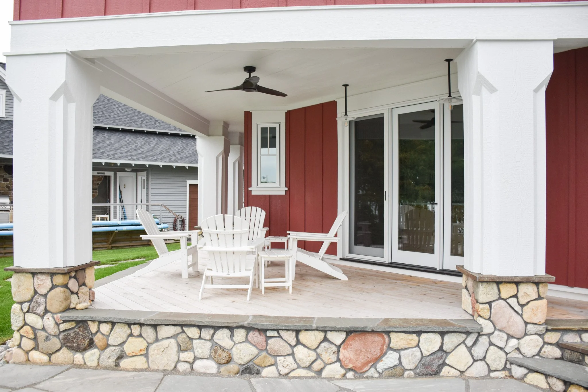 A covered outdoor patio with white chairs, a wooden floor, a ceiling fan, and a sliding glass door. The patio is supported by stone columns and is attached to a house with red and white siding.