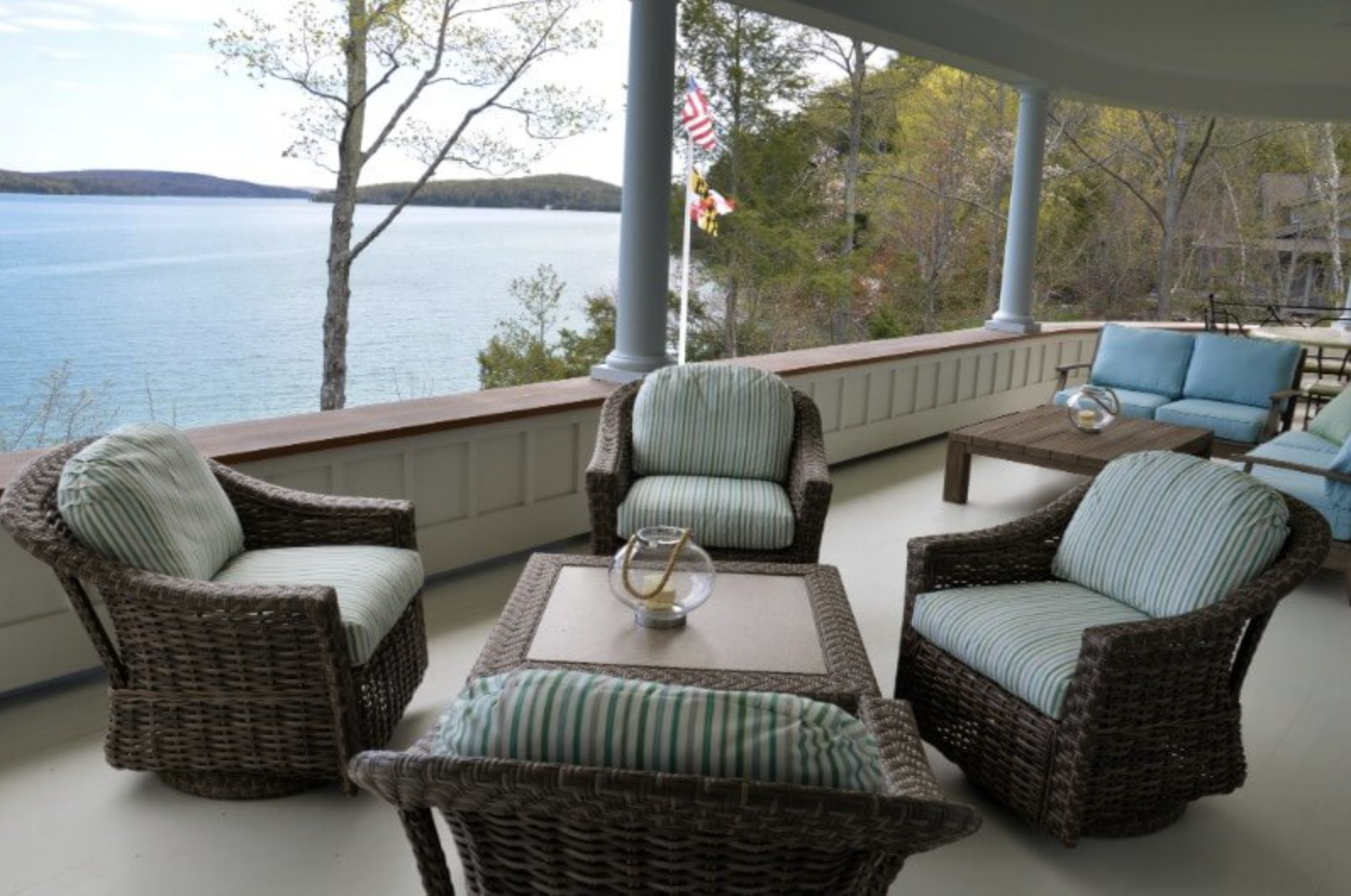 Porch with wicker and cushioned furniture overlooking a lake and trees, with American flags in the background.