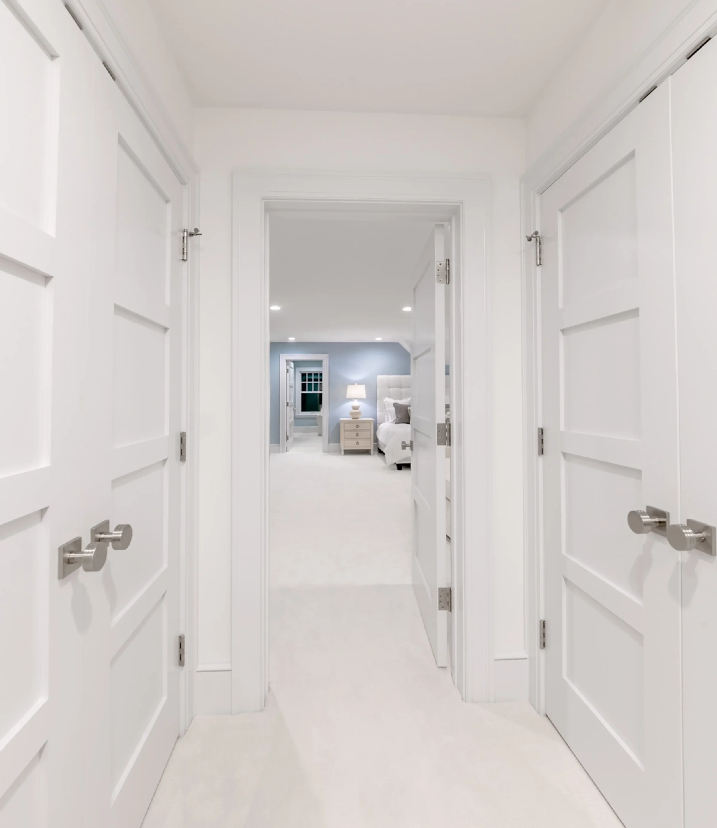 View of a bright, modern hallway leading to a bedroom with a nightstand, lamp, and bed visible in the background.