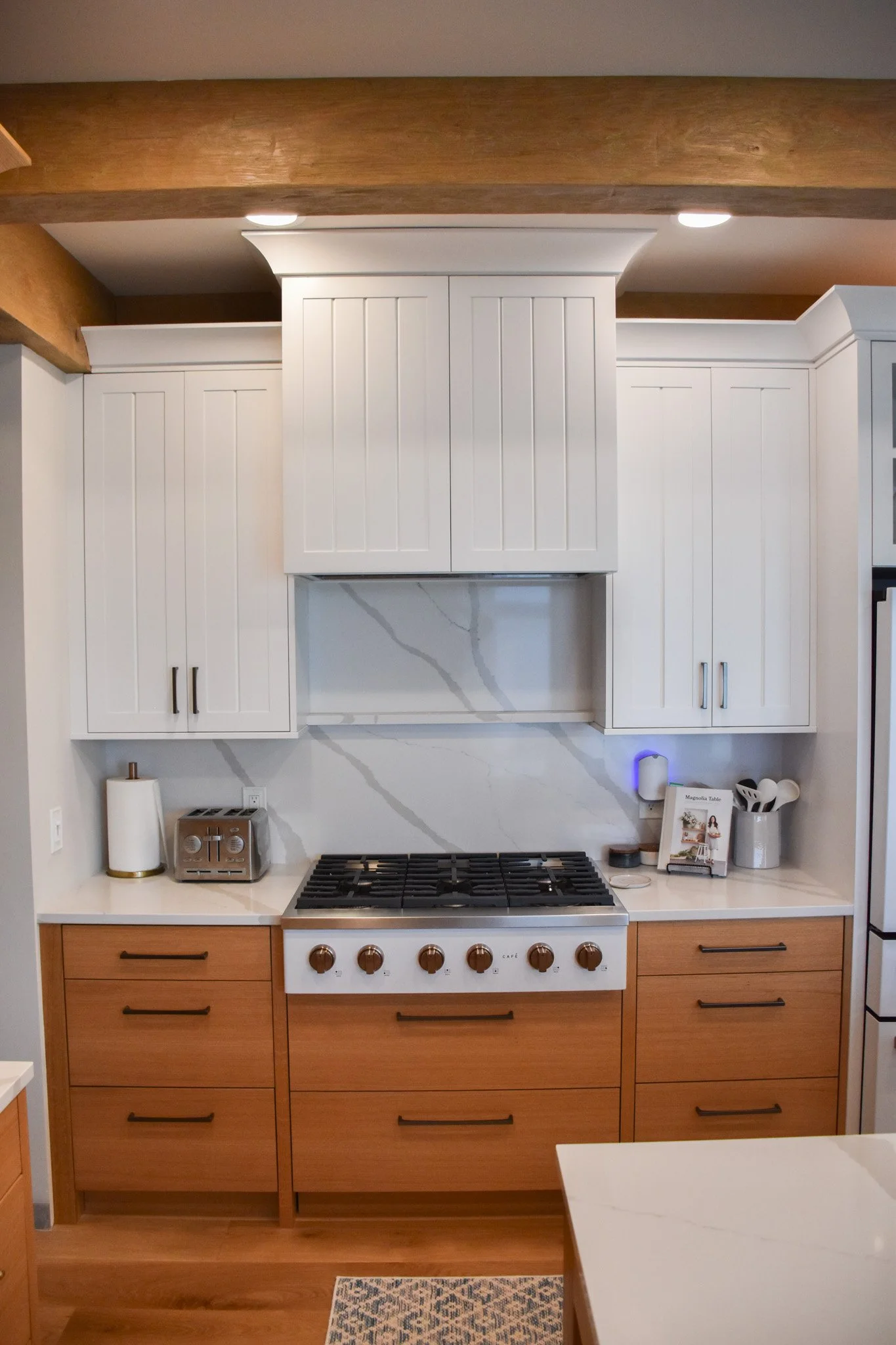 Kitchen with white upper cabinets, wooden lower cabinets, and a white stove with copper knobs. There is a paper towel roll, a toaster, and a small cookbook or magazine on the countertop. The backsplash is marble with gray veining, and a rug is on the