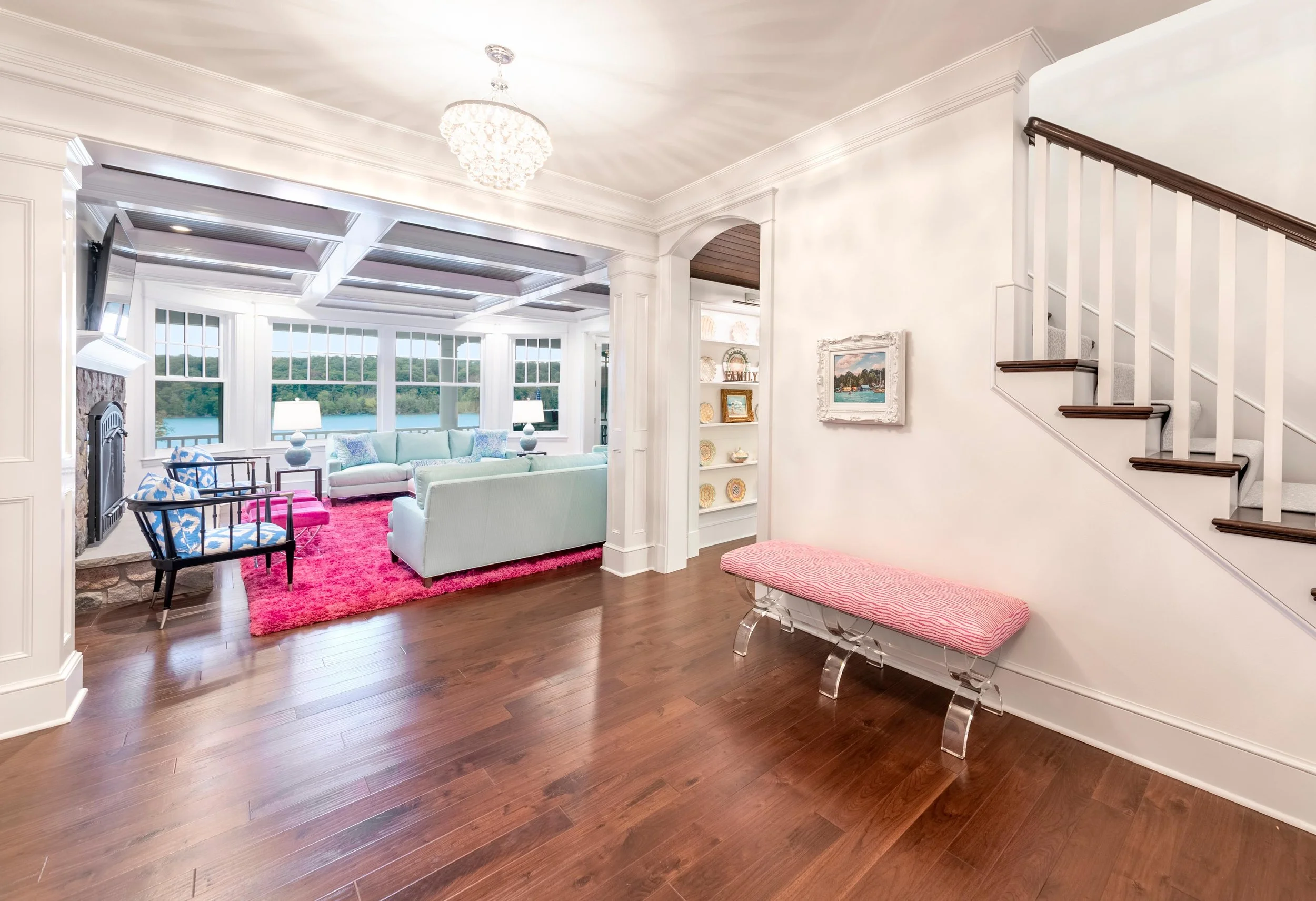 Interior of a bright living room with hardwood floors, large windows overlooking a lake, a stone fireplace, a pastel green sofa, colorful chairs, pink rug, and wall art.