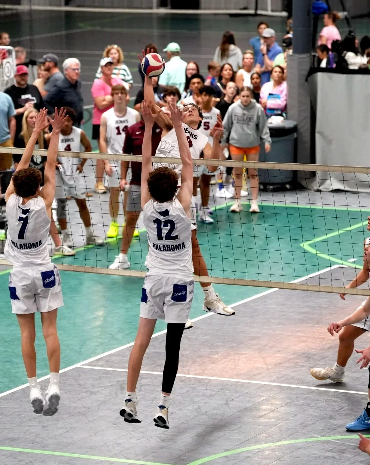 An indoor volleyball game with players from Oklahoma, one jumping to hit the ball over the net, while two players from the opposing team attempt to block.