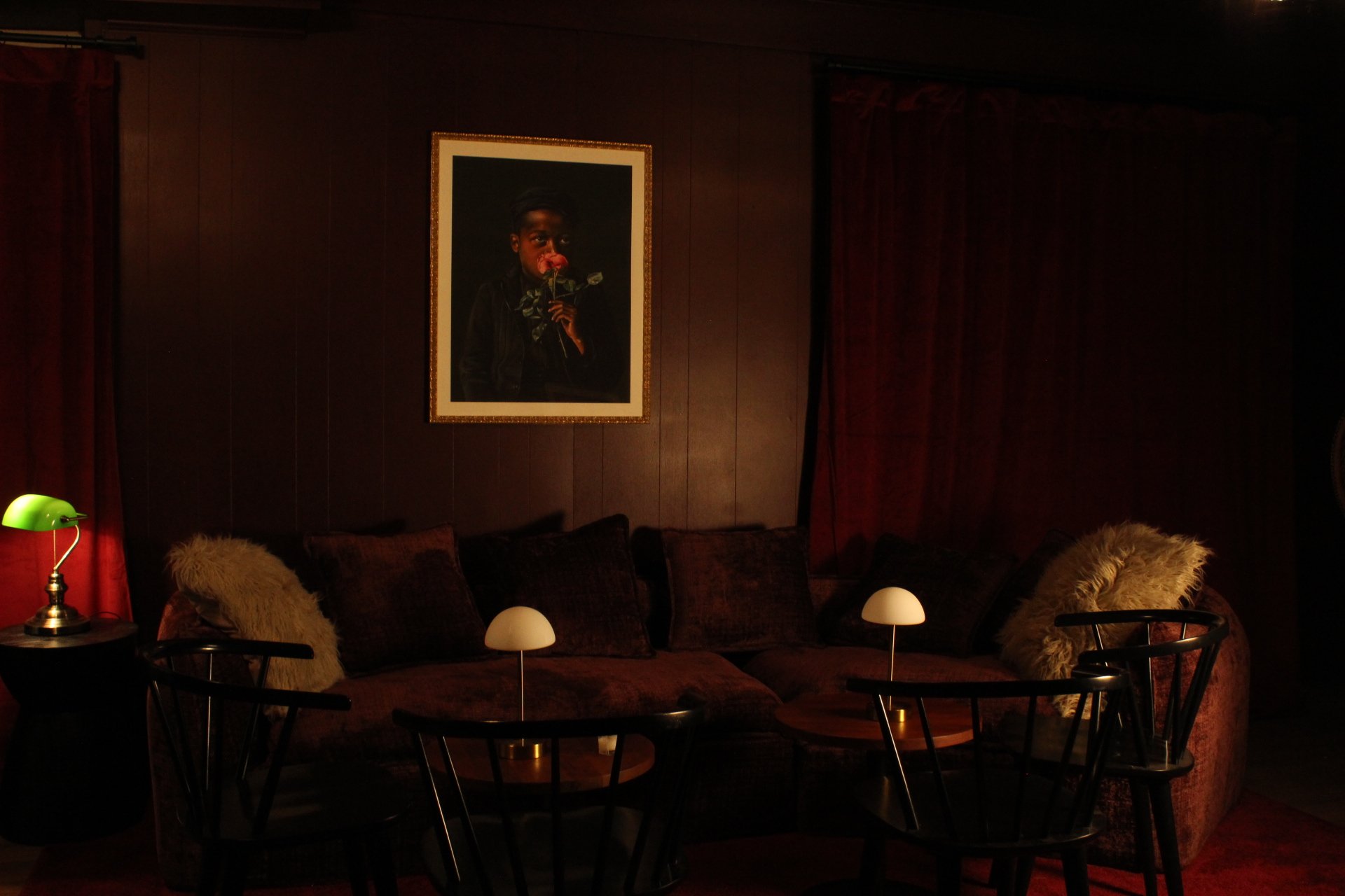 A dimly lit living room with a dark wooden wall, a framed portrait of a person holding a rose, brown patterned sofa with fuzzy pillows, a green desk lamp, and small white table lamps on side tables.